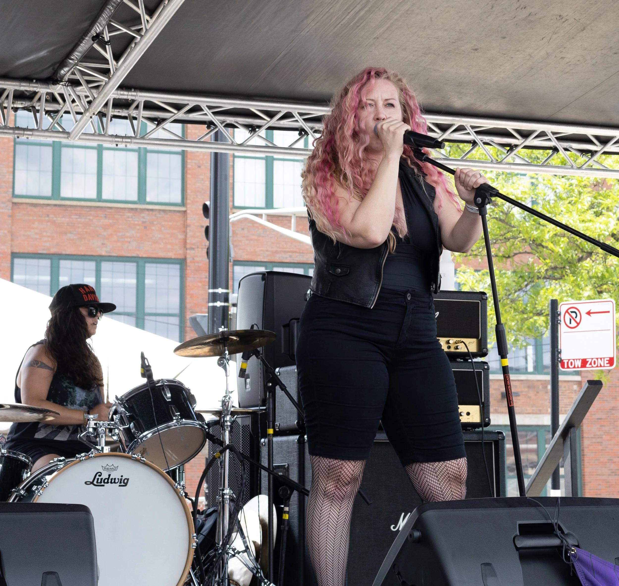 A female singer with pink curly hair performing on an outdoor stage with a microphone, wearing fishnet stockings and black clothing, backed by a drummer playing a Ludwig drum set.