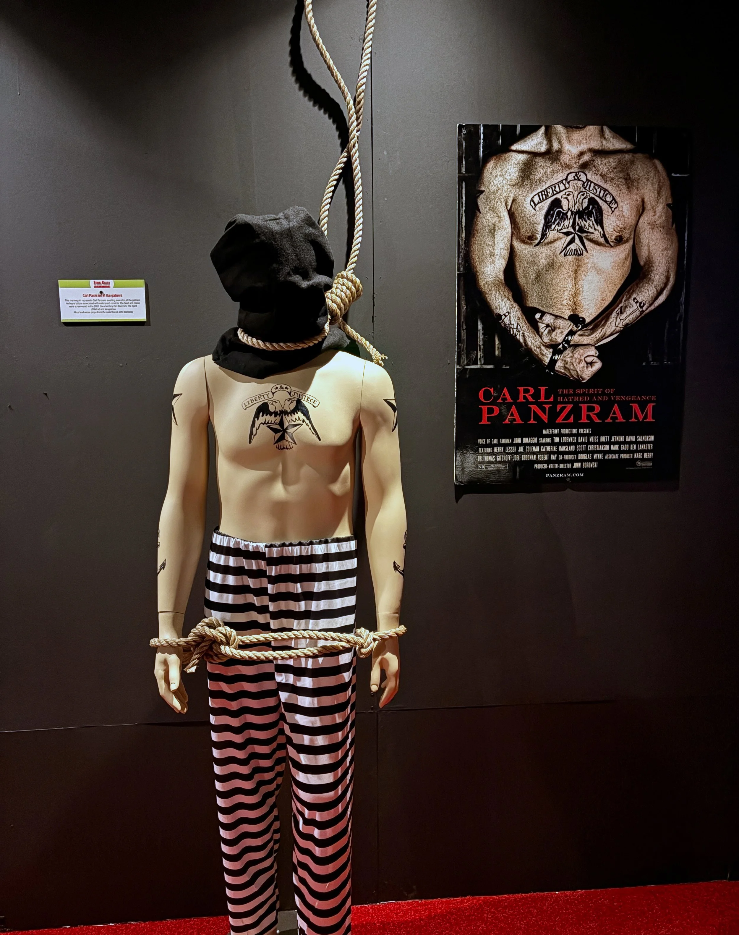 A mannequin with a tattoo of an eagle and the words "Liberty & Justice" on its chest, wearing black and white striped pants, standing in front of a dark wall and a poster of Carl Panzram. The mannequin's head is covered with a gray hoodie, and it is 