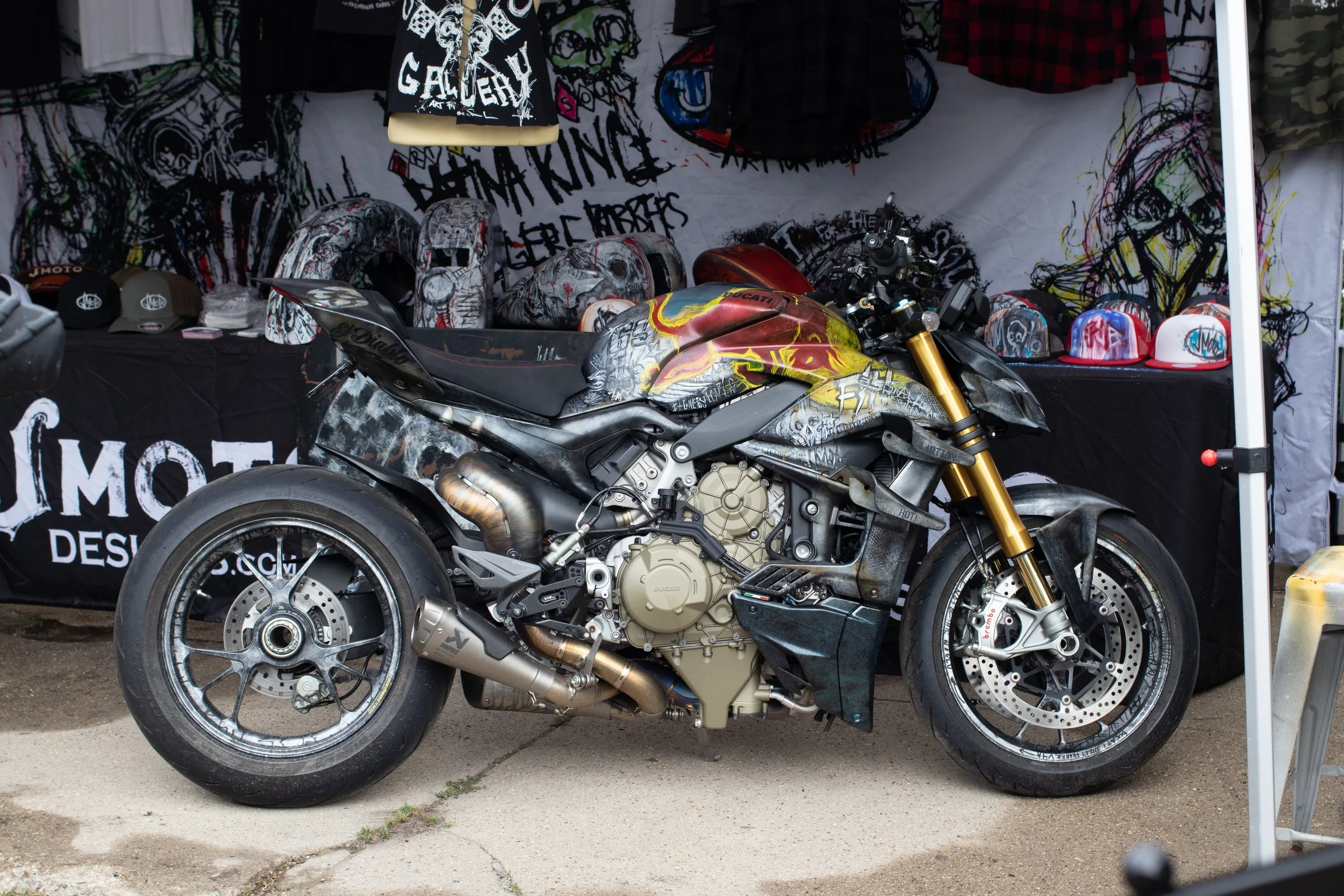 A motorcycle with custom graffiti-style paintwork displayed in front of a booth. The booth has helmets, clothing, and hats with similar artistic designs. There is a large graffiti-style banner behind the booth.