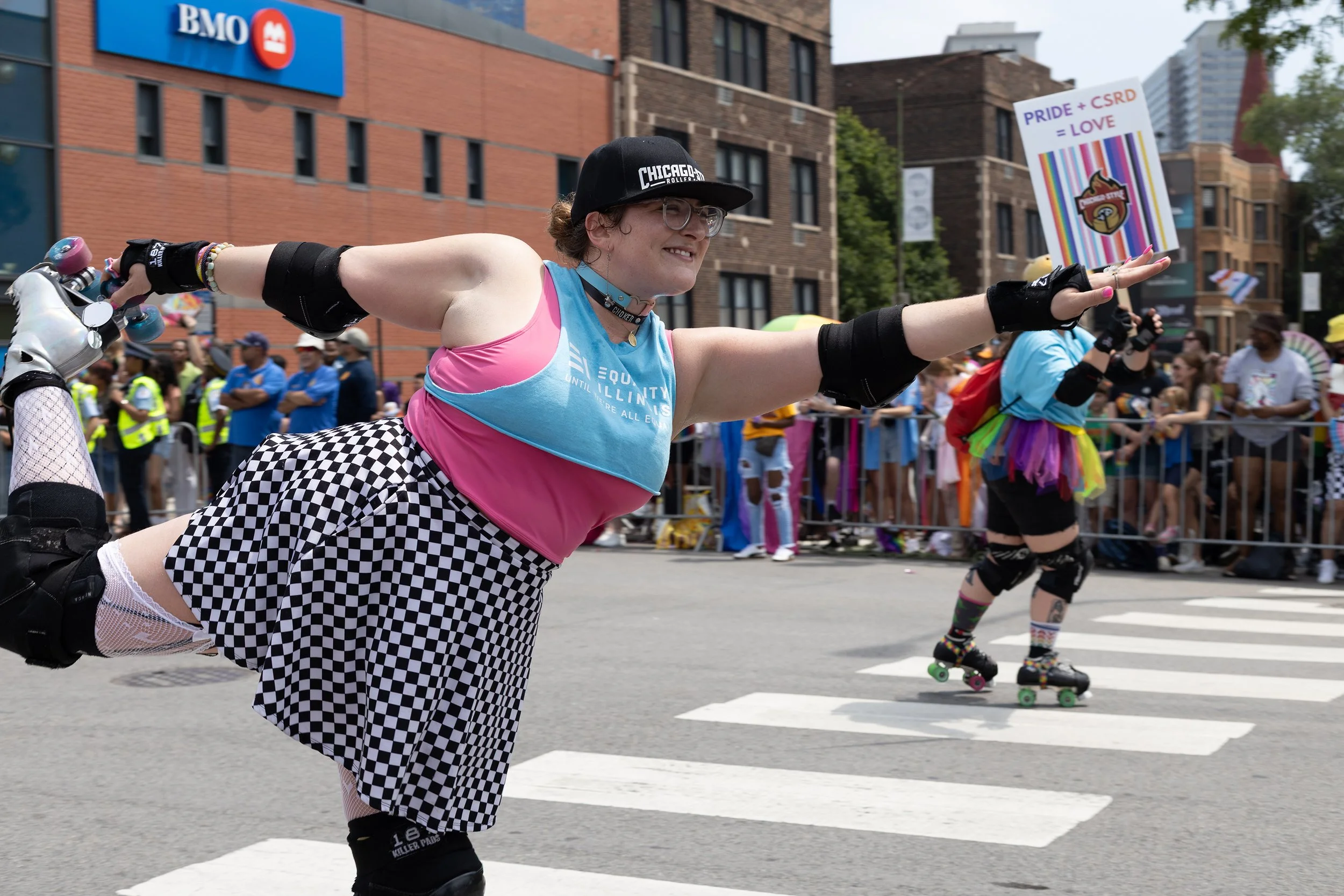 A person in colorful roller derby skates, wearing a black cap, glasses, a pink and blue tank top, and checkered shorts, balances on one leg with arms outstretched during a parade on a city street. Background includes other skaters, spectators, and bu