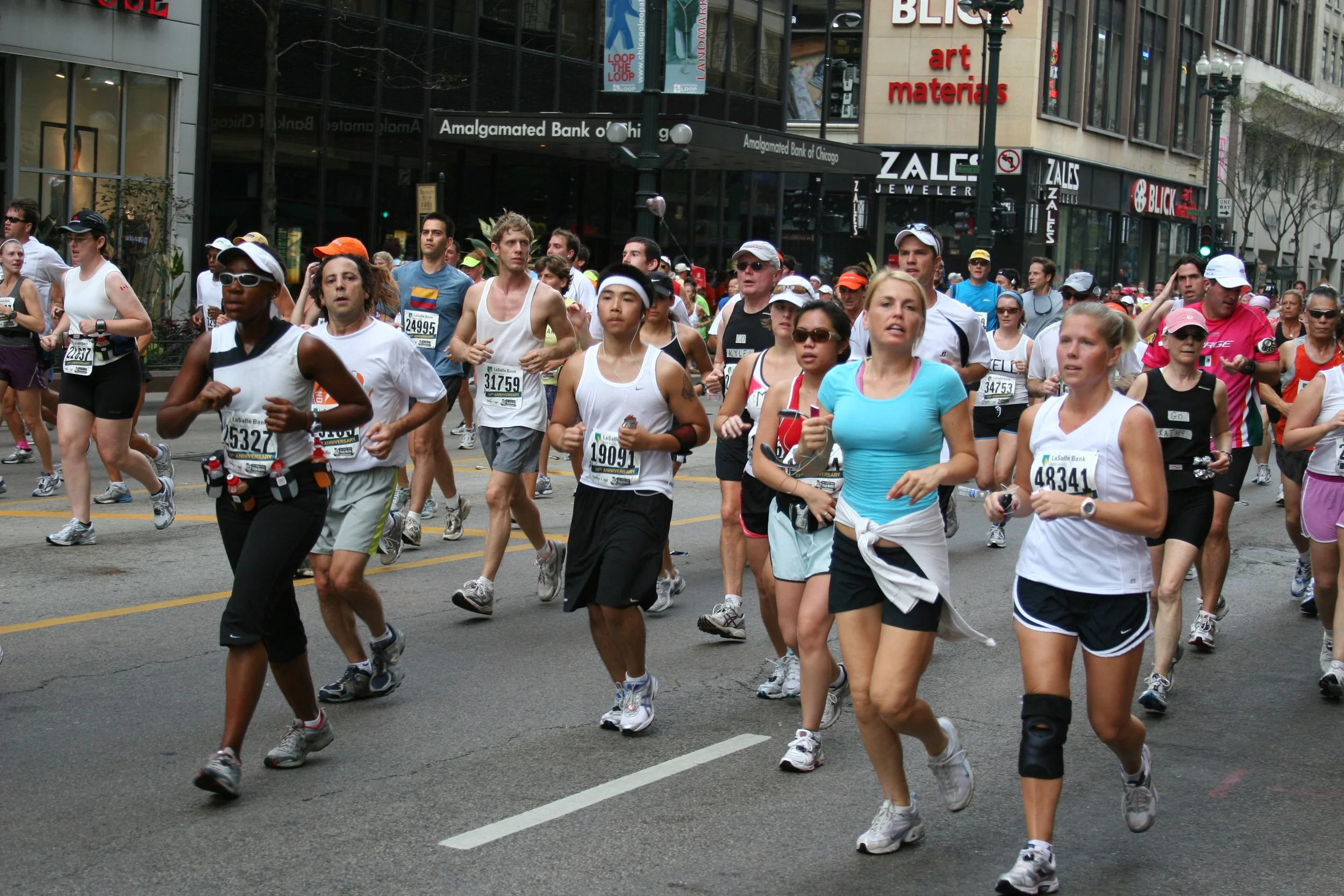 A group of marathon runners participating in a race on a city street, with buildings and storefronts in the background.