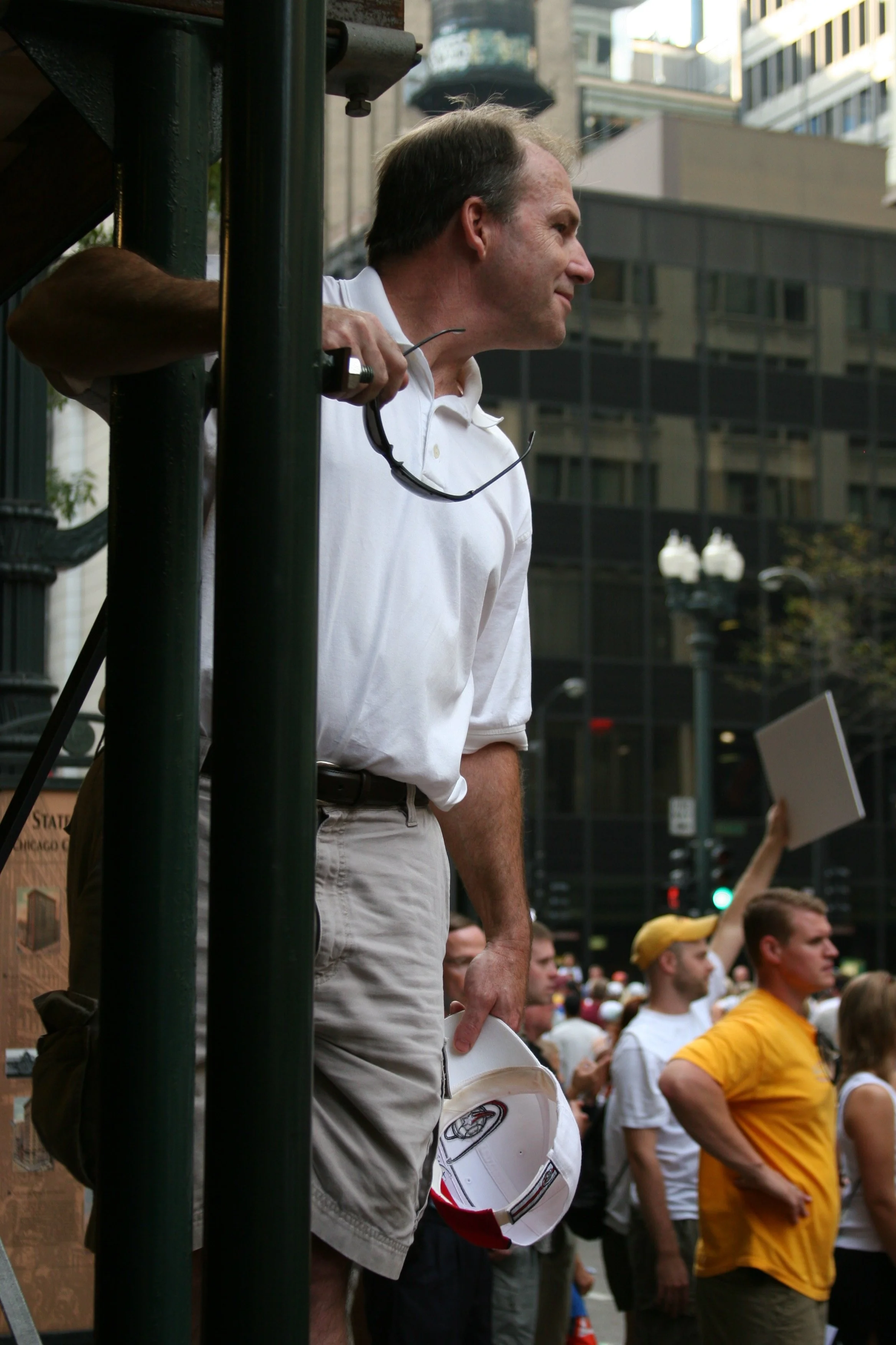 A man with a white shirt holding a helmet, surrounded by a crowd of people, some wearing yellow and white shirts, in an urban setting with tall buildings.