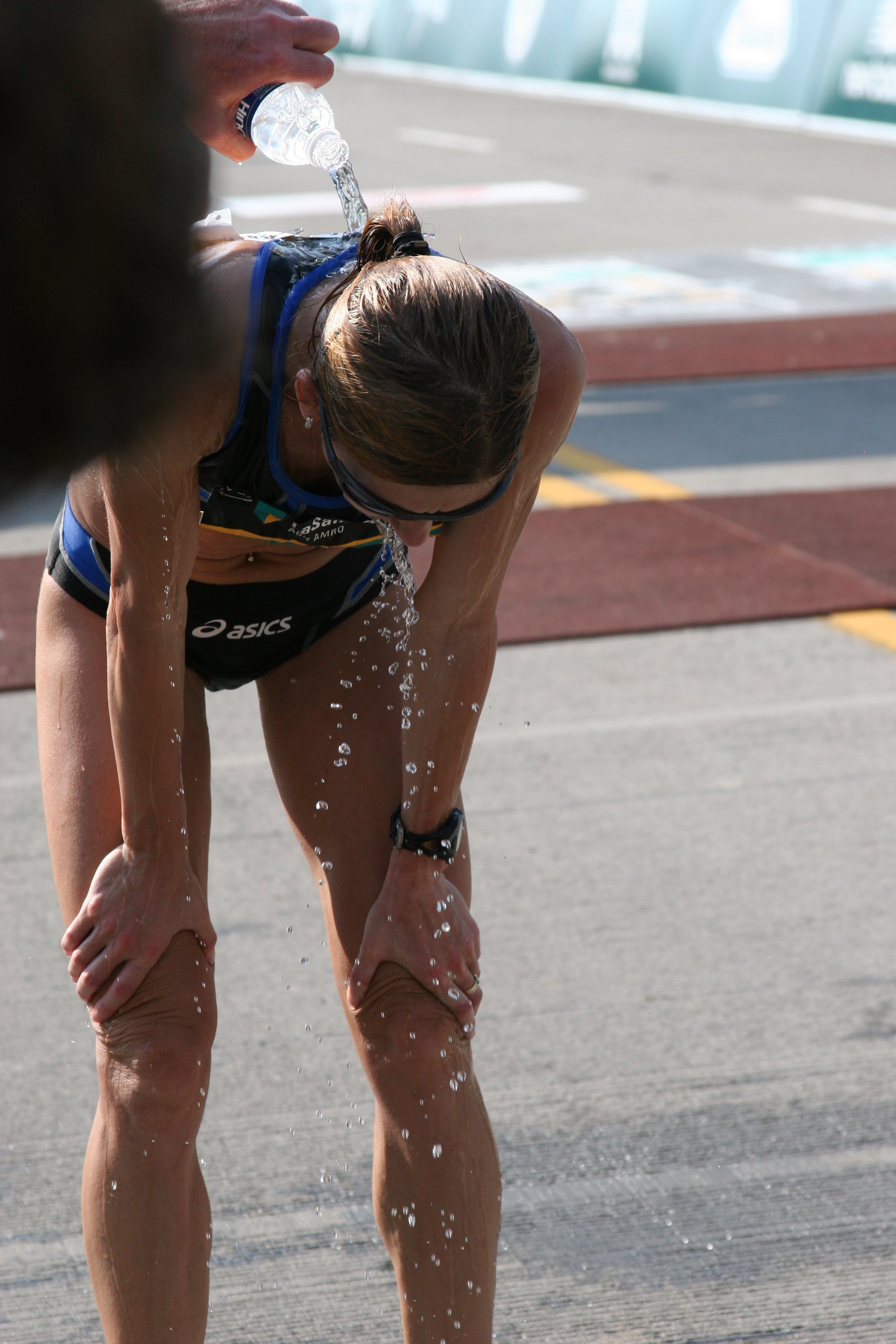 A female athlete, wearing sunglasses and athletic gear, is bent over and pouring water from a plastic bottle over her head, likely after a race or workout, on a sunny outdoor track.