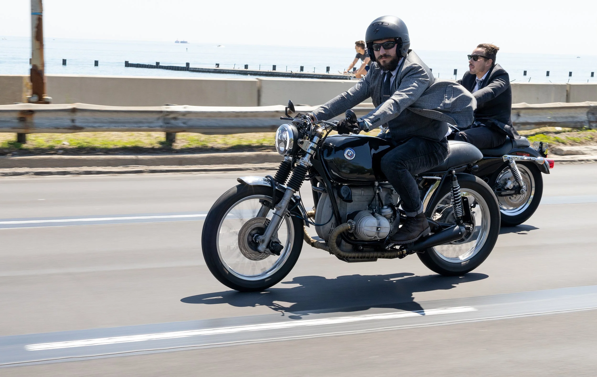 Two men in suits riding a motorcycle along a coastal road, with the ocean and a pier in the background, one man wearing a helmet and sunglasses.