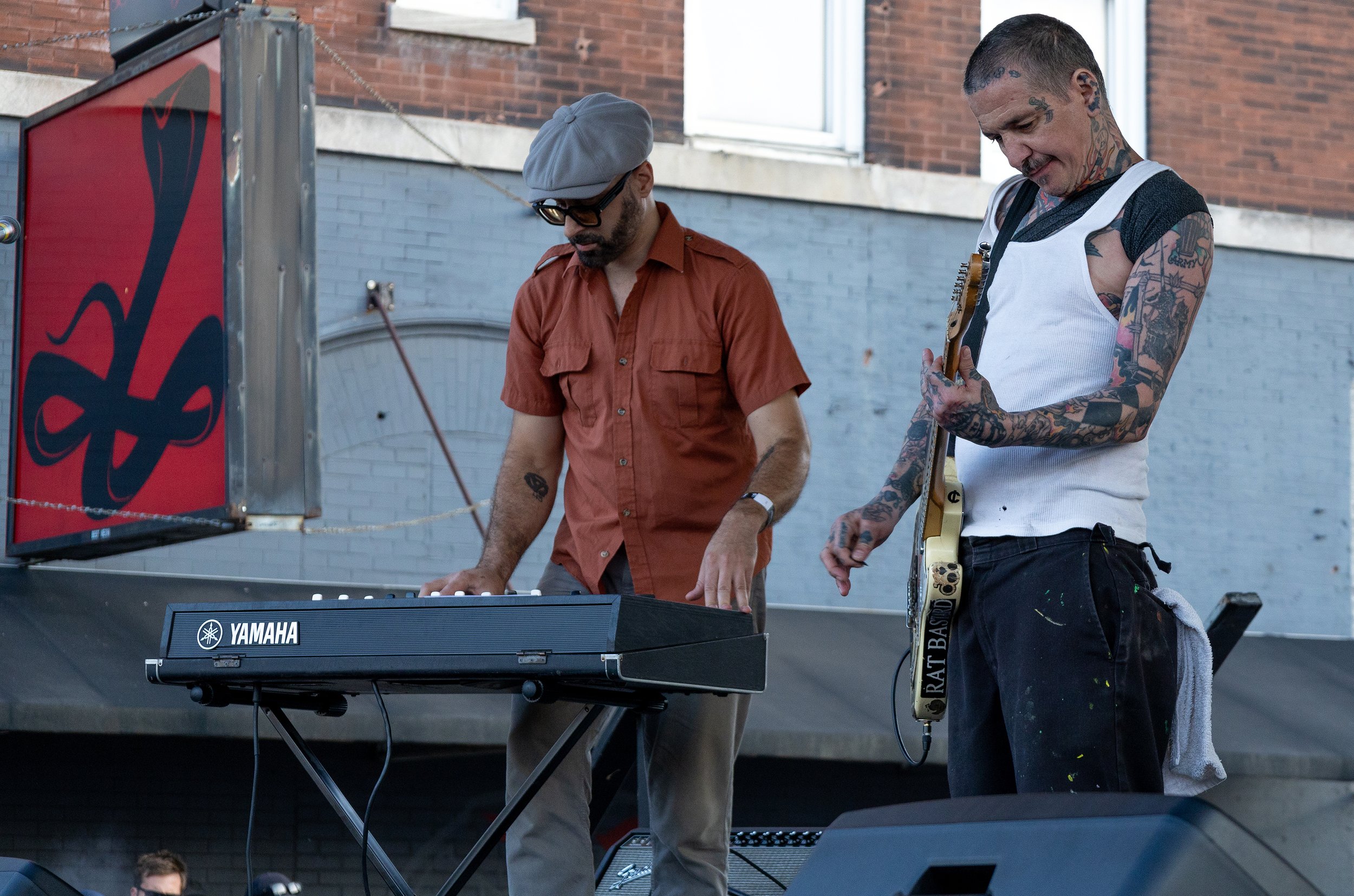 Two musicians perform outdoors near a brick building. One is playing a Yamaha keyboard, and the other is holding a guitar with tattoos on his arms, wearing a white tank top, and looking down.
