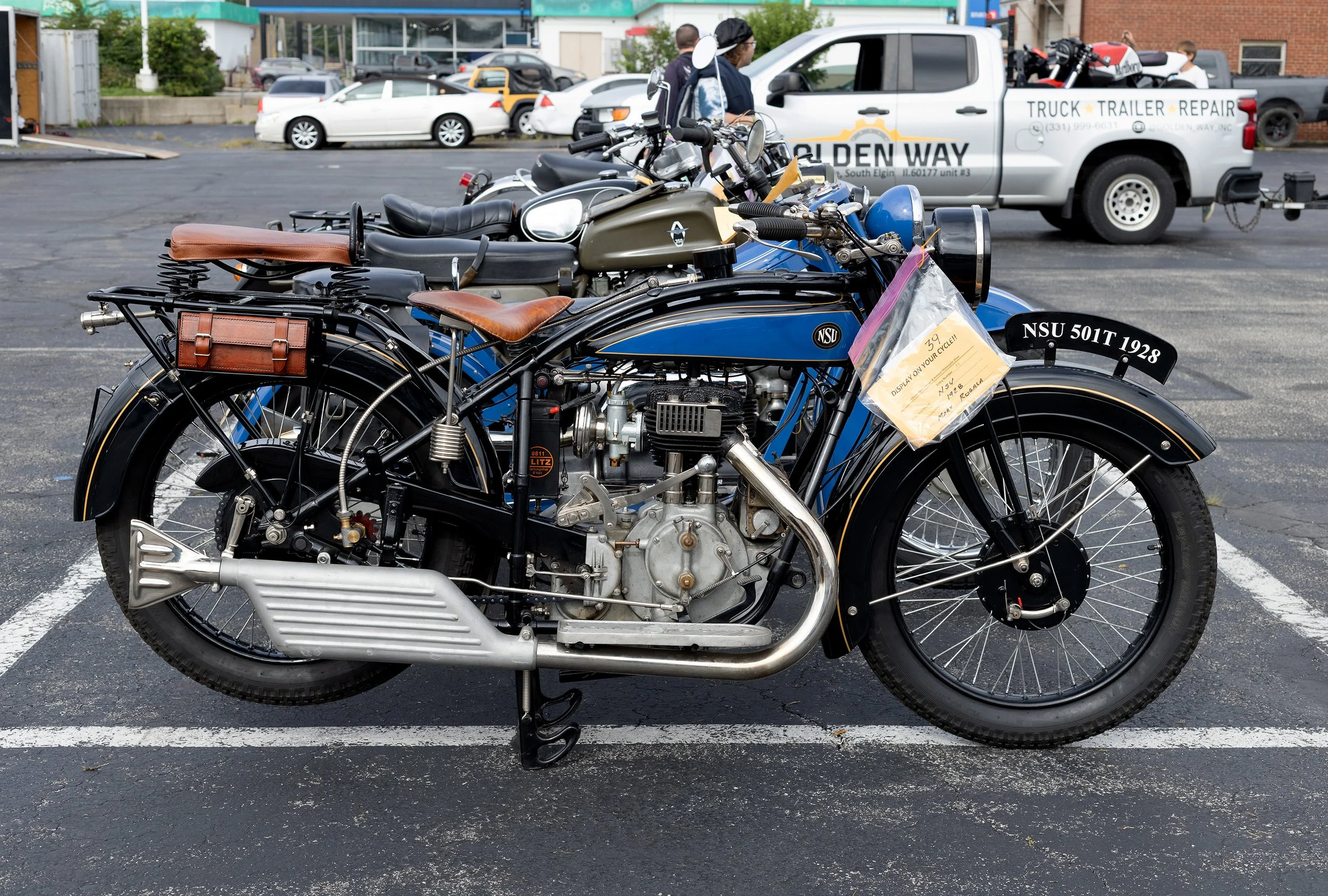A vintage motorcycle with a blue and black color scheme, brown leather seat, and a small leather saddlebag, parked in a parking lot.