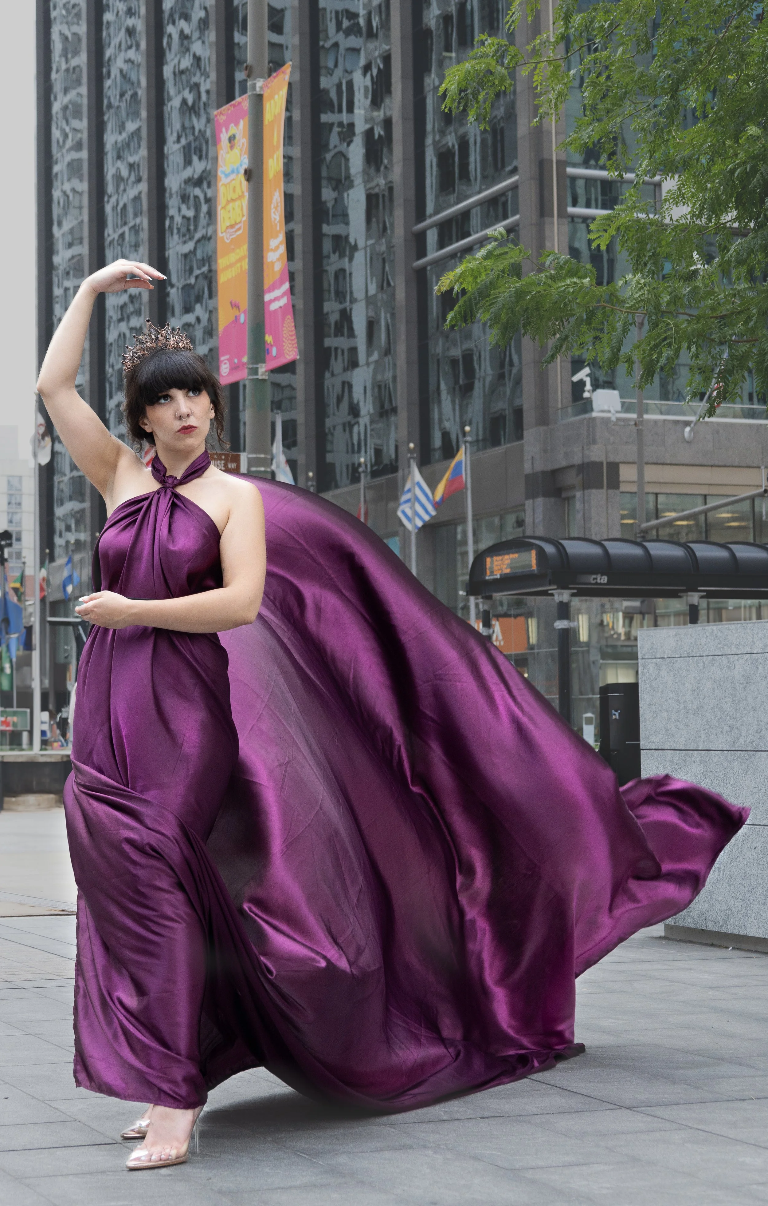 A woman in a flowing purple gown and clear high heels is dancing or posing in an urban setting with modern glass buildings and flags in the background. She is wearing a crown and has dark hair with bangs.