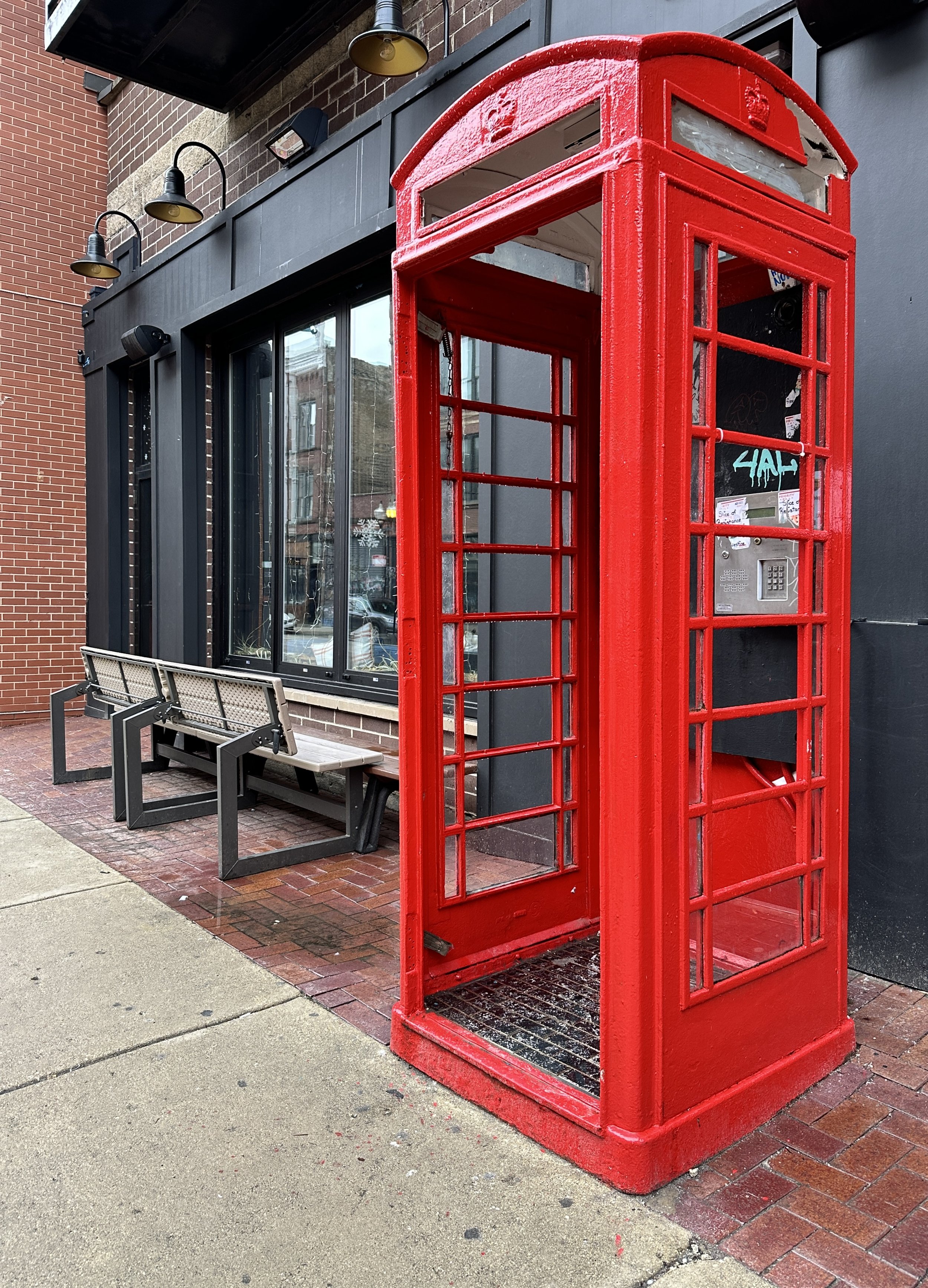 Red telephone booth on a city sidewalk, next to a bench and a building with large windows and modern black exterior.