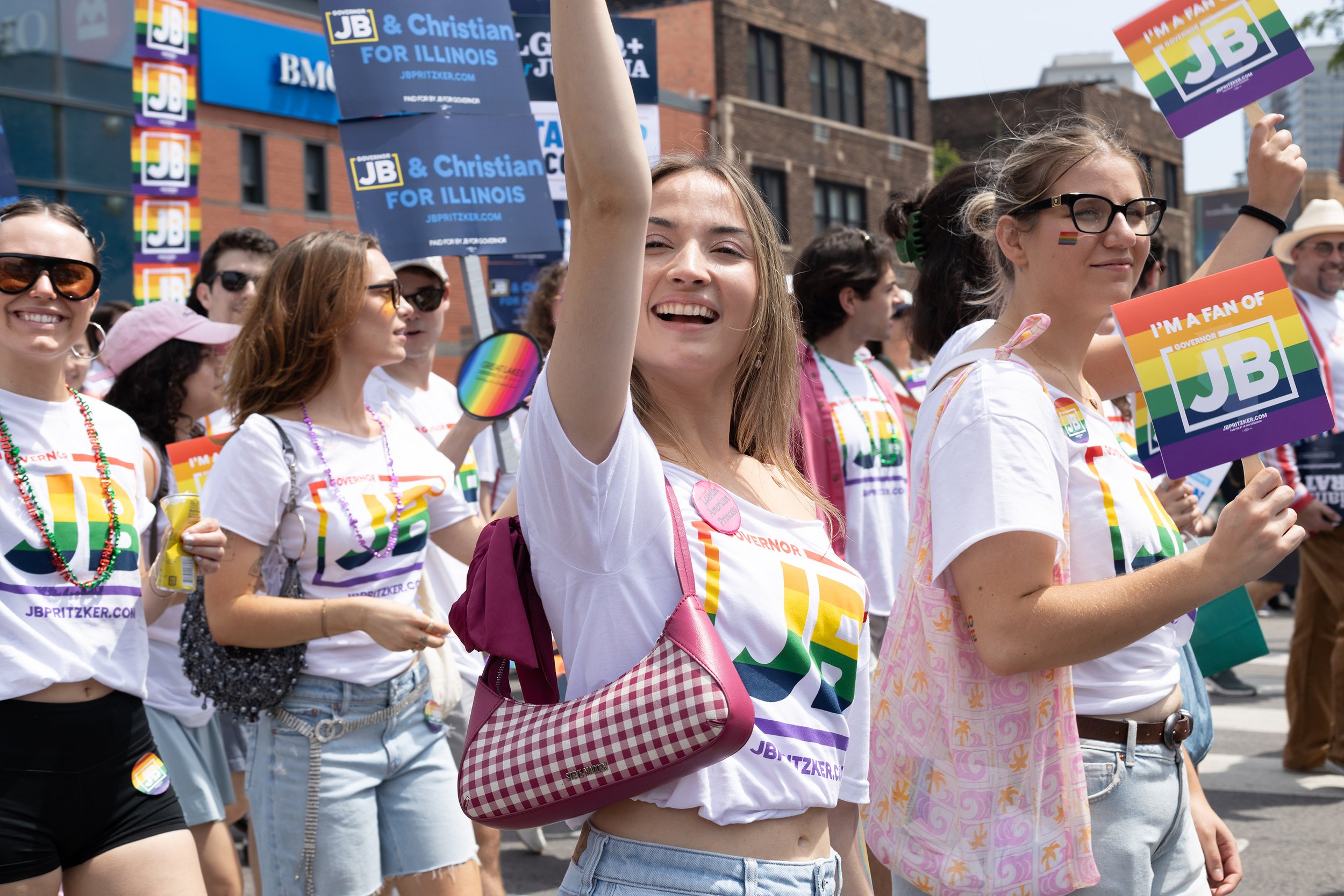 Group of people participating in a pride parade, holding rainbow-colored signs and wearing rainbow-themed accessories, with city buildings in the background.