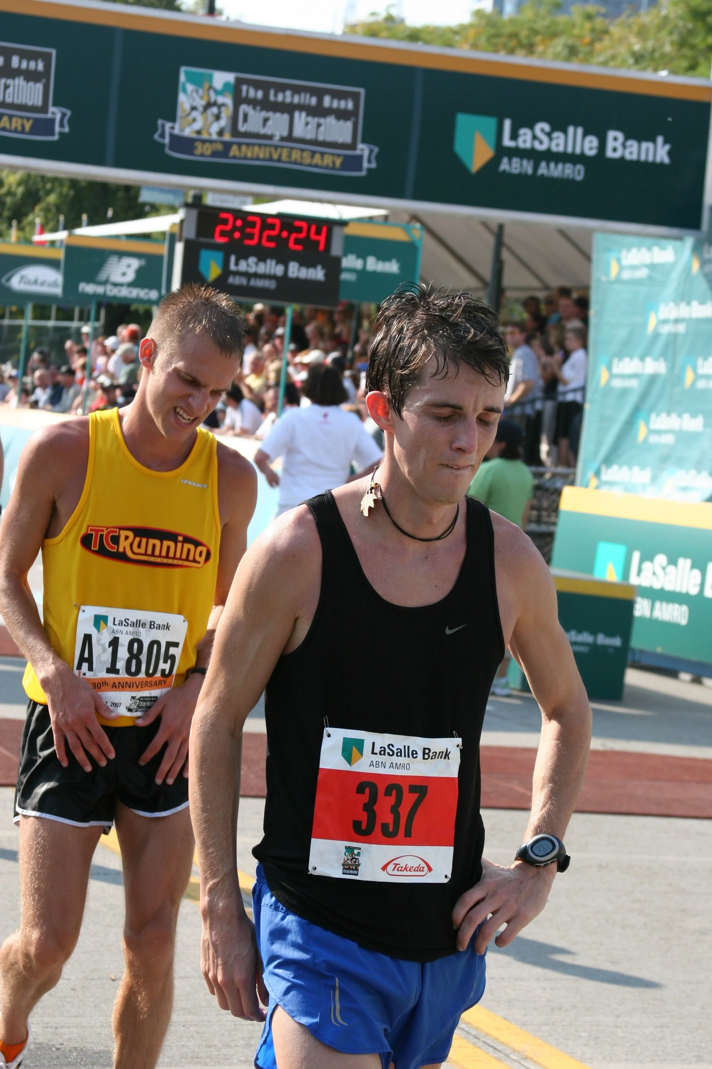 Two male marathon runners at the finish line, one wearing a yellow TCRunning tank top and the other in a black sleeveless shirt, with race bibs and a digital watch, under banners and a crowd of spectators.