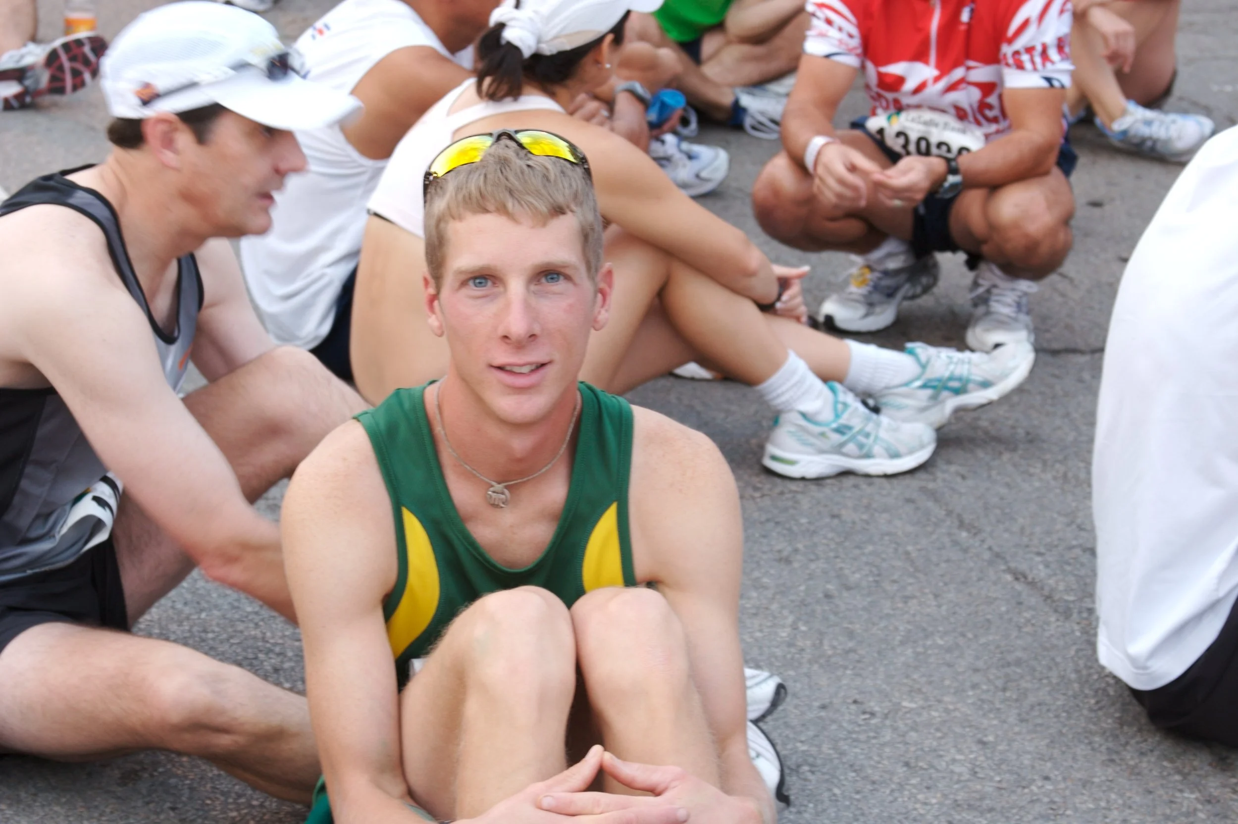 A group of people sitting on the ground, many wearing athletic clothing and bibs, with a young man with blue eyes in the foreground wearing a green and yellow athletic shirt. Some are wearing white caps and sunglasses, and they appear to be resting a
