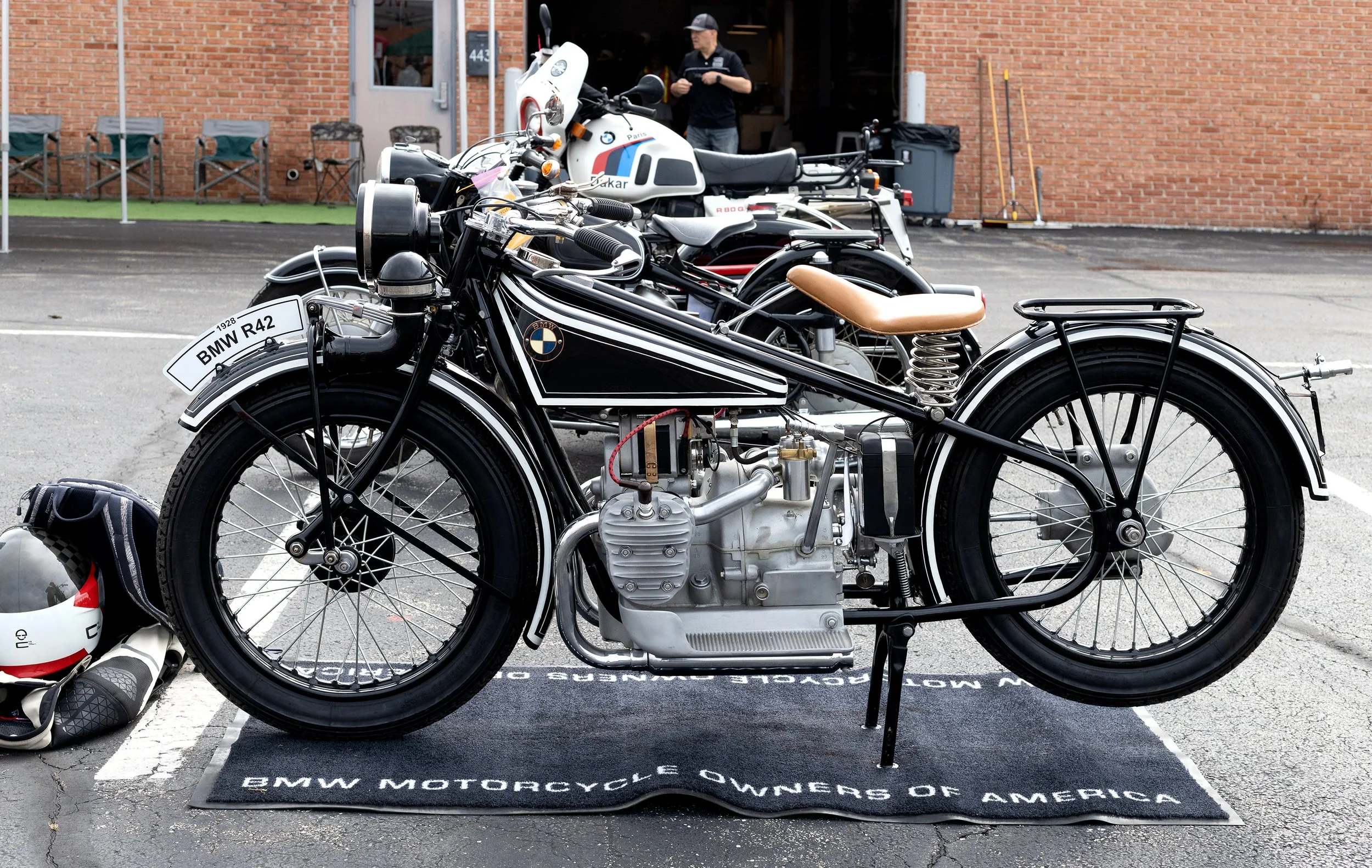 A vintage BMW motorcycle on display at a BMW motorcycle event, with a white and black color scheme, a leather seat, and a license plate that reads 'BMW R42'. A helmet and riding gear are placed on the ground nearby. In the background, a person is sta