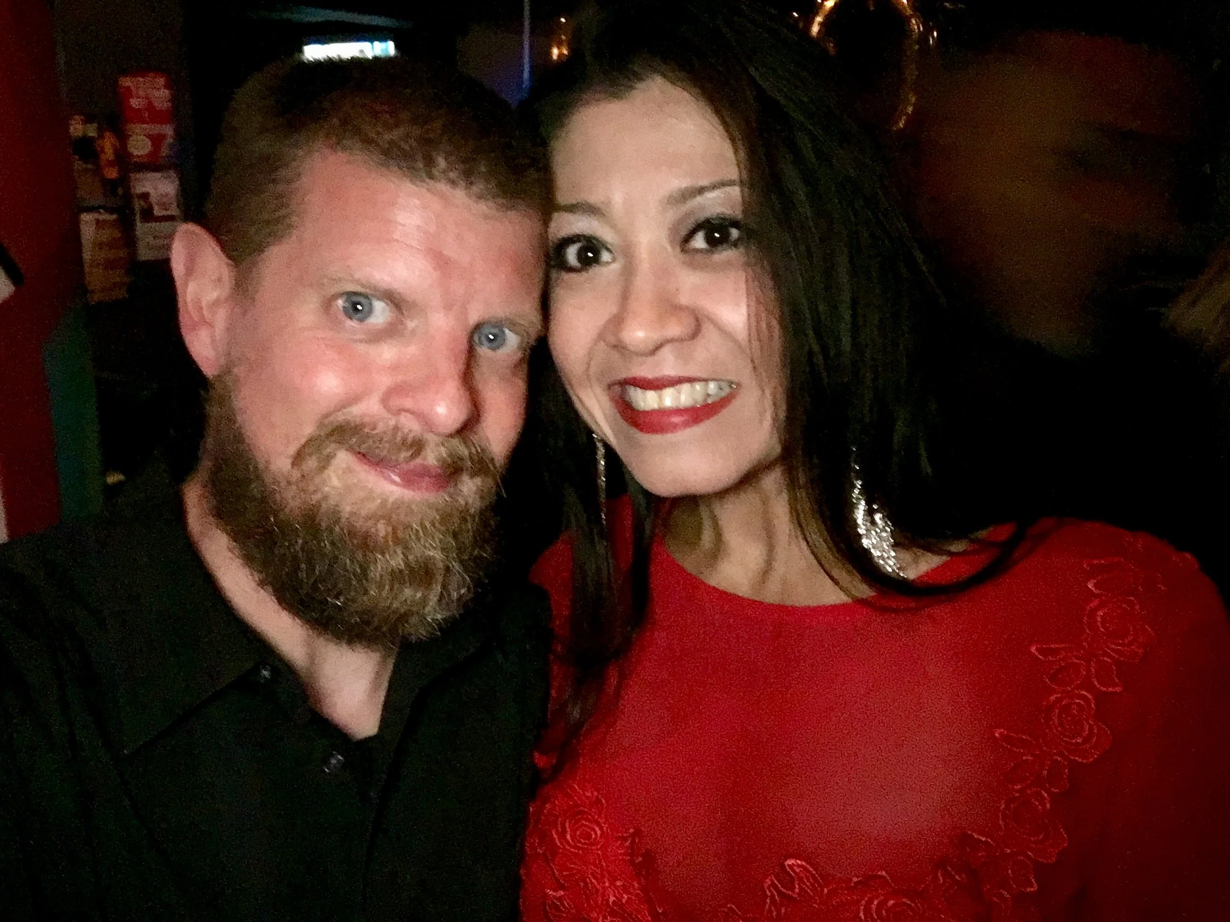 A man with a beard and a woman with dark hair and red lipstick posing closely together for a photo in a dark setting.