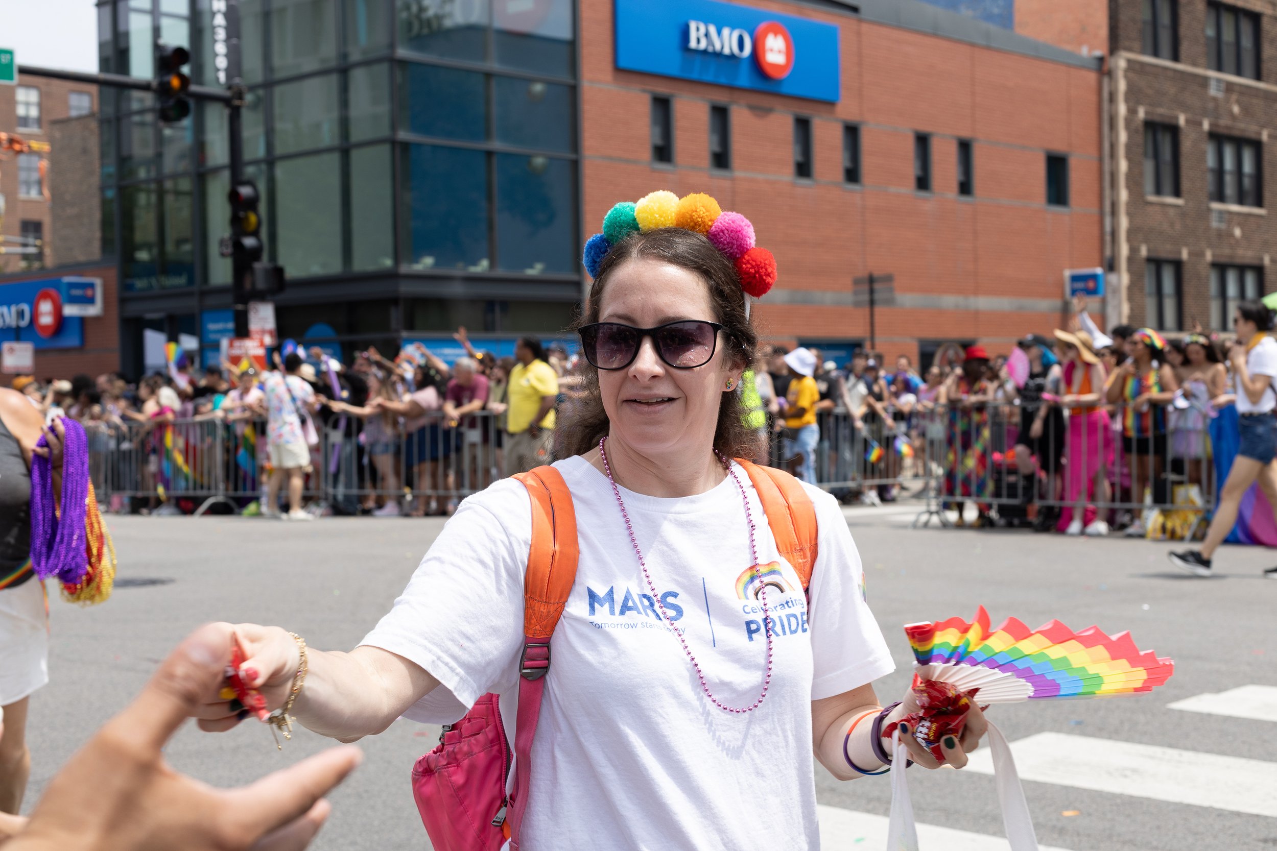 A woman wearing sunglasses and a rainbow pom-pom headband during a Pride parade, holding a rainbow-colored fan and pink backpack, with people and a building with a BMO sign in the background.