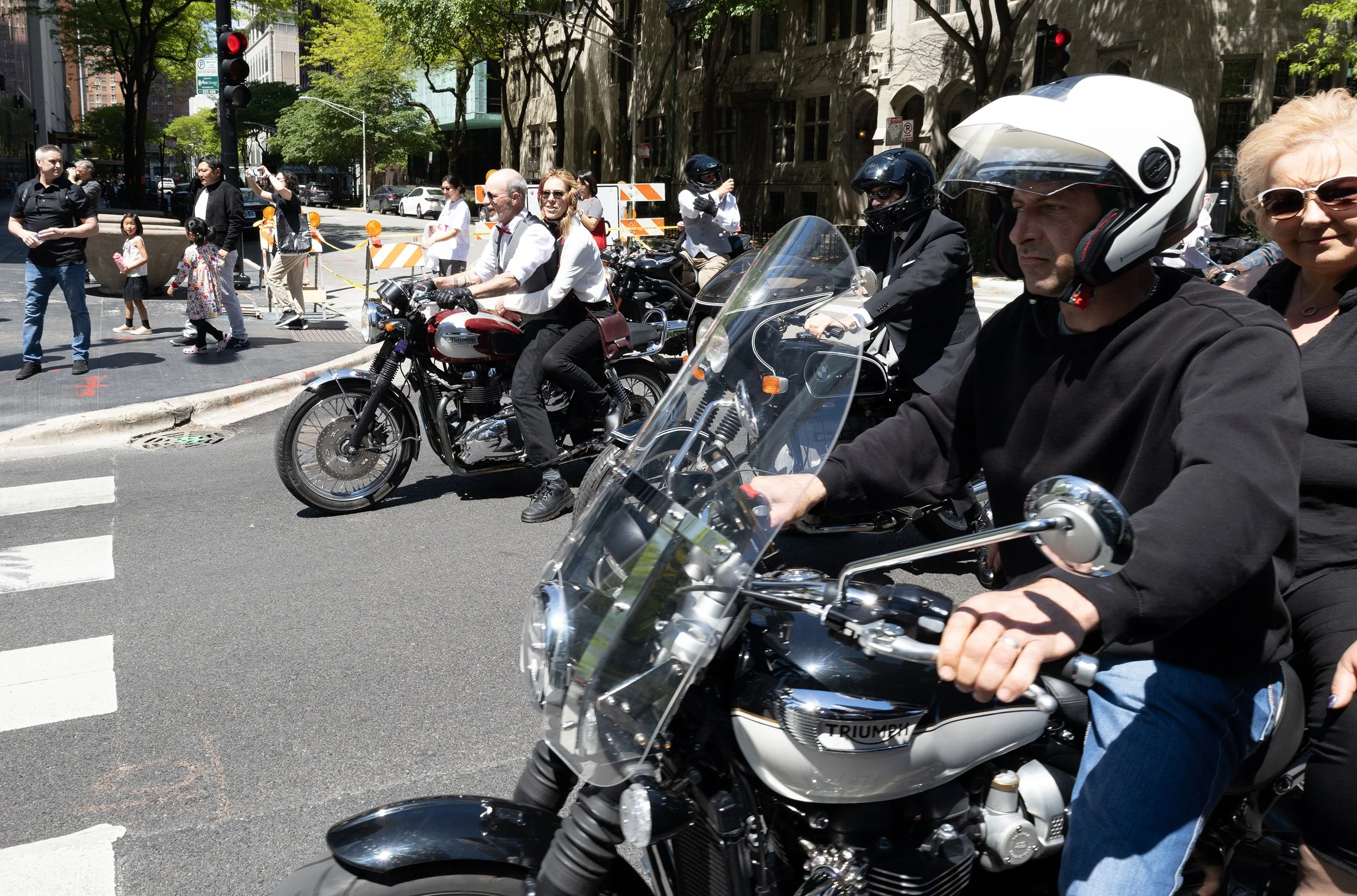 People crossing the street at a busy urban intersection, with some on motorcycles, others walking. The scene shows a mixed crowd with urban buildings and trees in the background.
