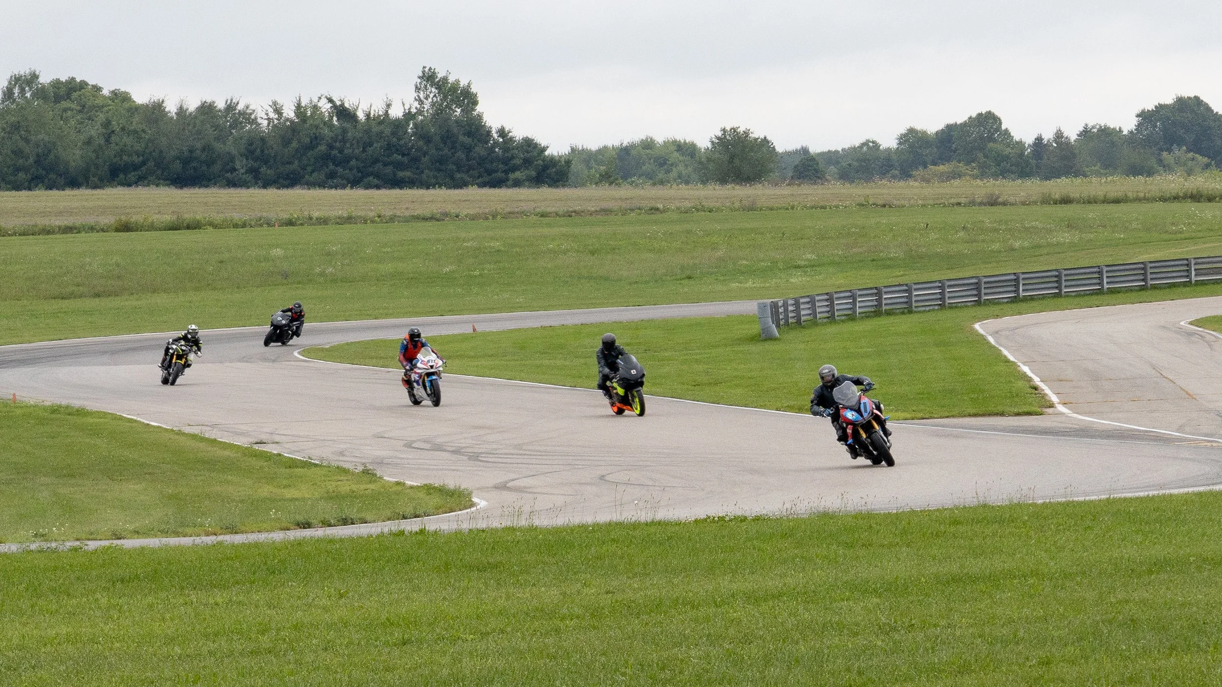 Multiple motorcyclists riding on a curved race track surrounded by green grass and trees in the background on an overcast day.