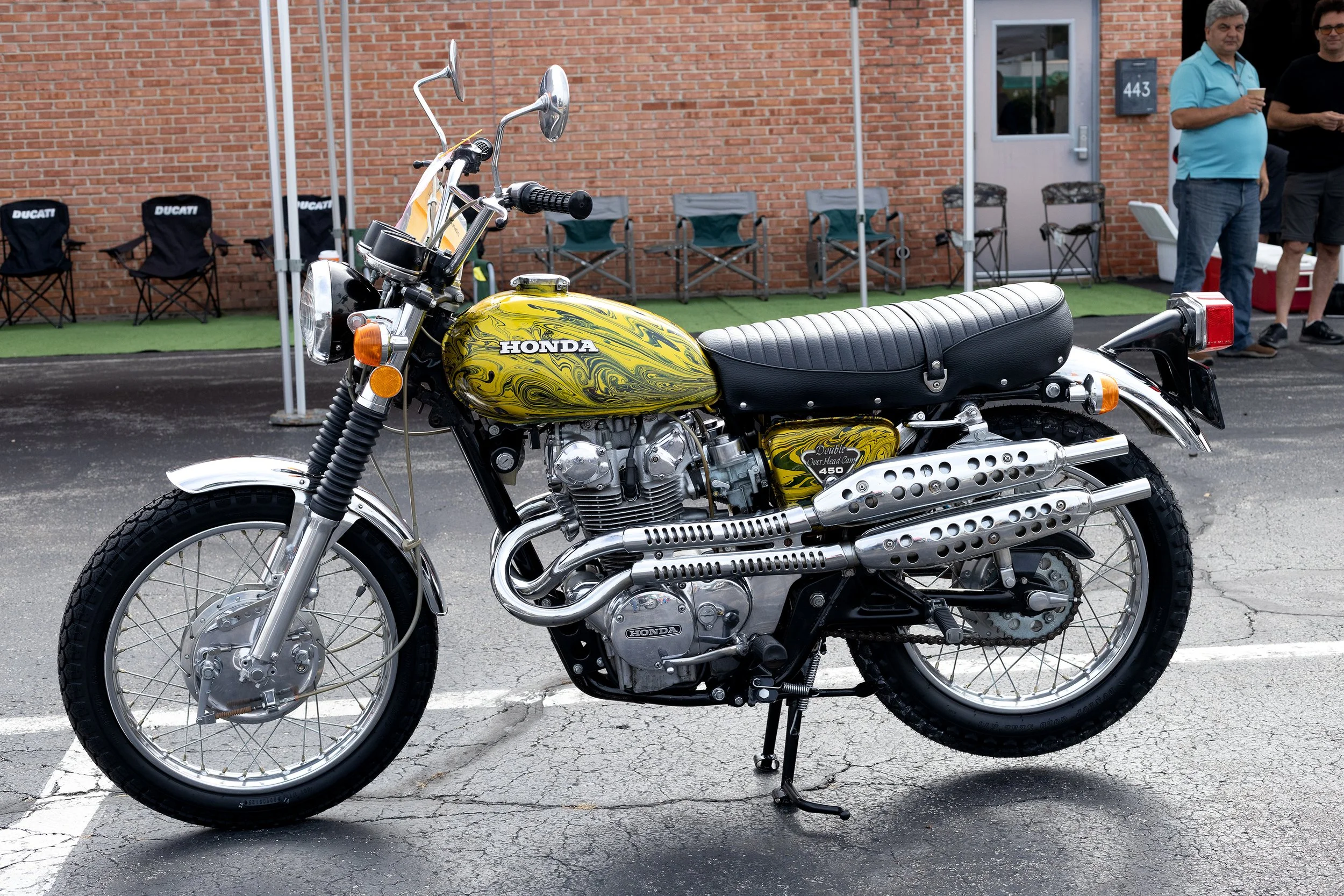 A vintage yellow Honda motorcycle with a black seat parked in front of a brick building, with people standing nearby.