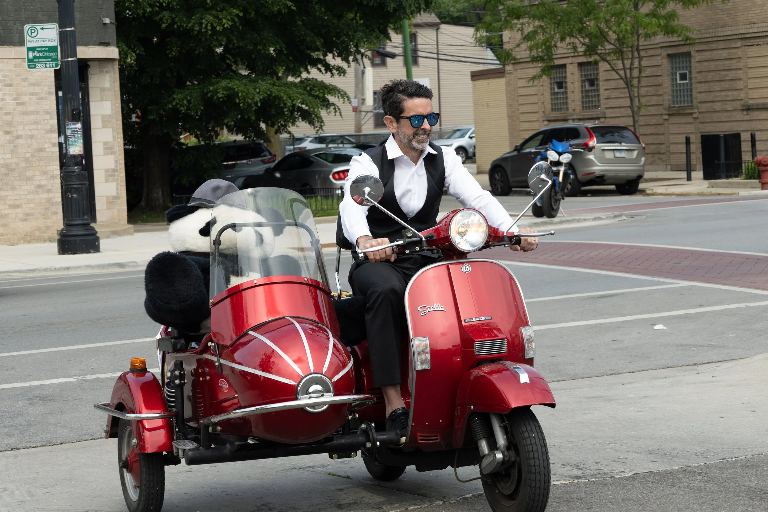 A man with dark hair, wearing blue sunglasses, a white shirt, and black vest, riding a red three-wheeled scooter with a sidecar, on a city street. There are parked cars, trees, and buildings in the background.