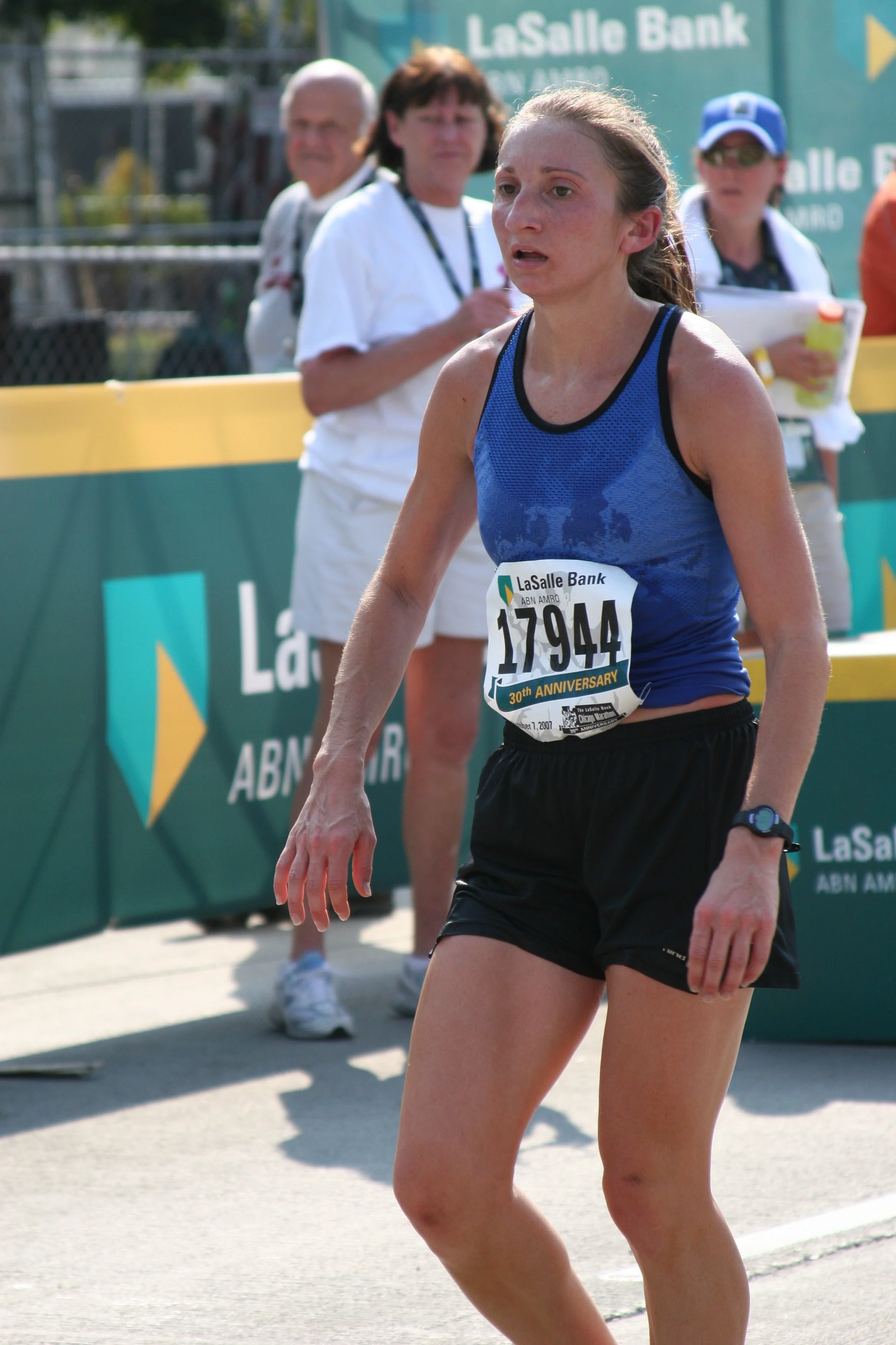 A female runner in a blue sports tank top and black shorts participating in a marathon, with a race bib number 17944, near a green and yellow LaSalle Bank event banner. Two women and a man are in the background, one of the women holding a notepad and