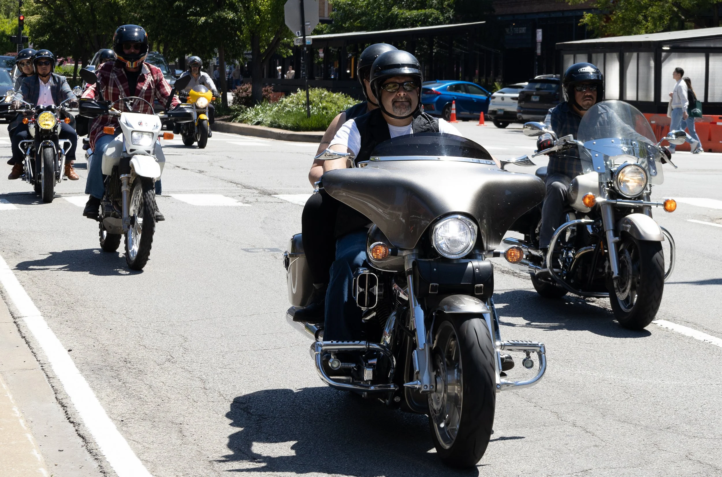 A group of motorcyclists riding on a city street with cars parked nearby and pedestrians walking on the sidewalk.