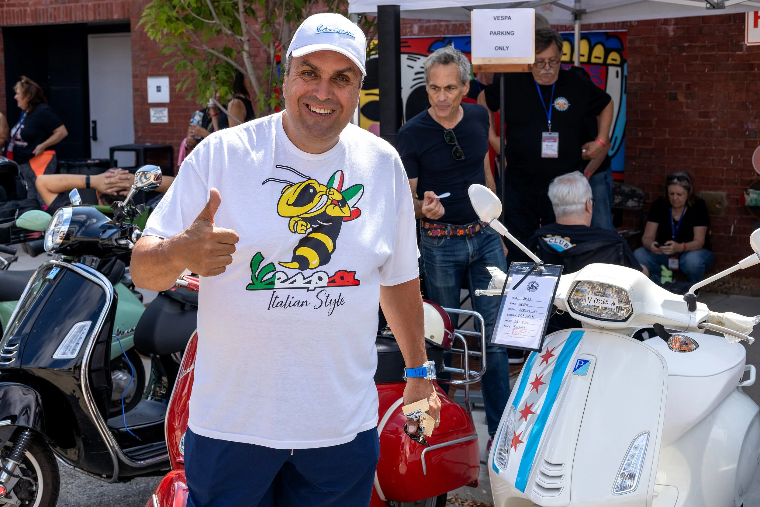 Man smiling and giving a thumbs-up at a scooter exhibit during a Vespa event, with other people in the background, some looking at scooters and one sitting on a scooter.