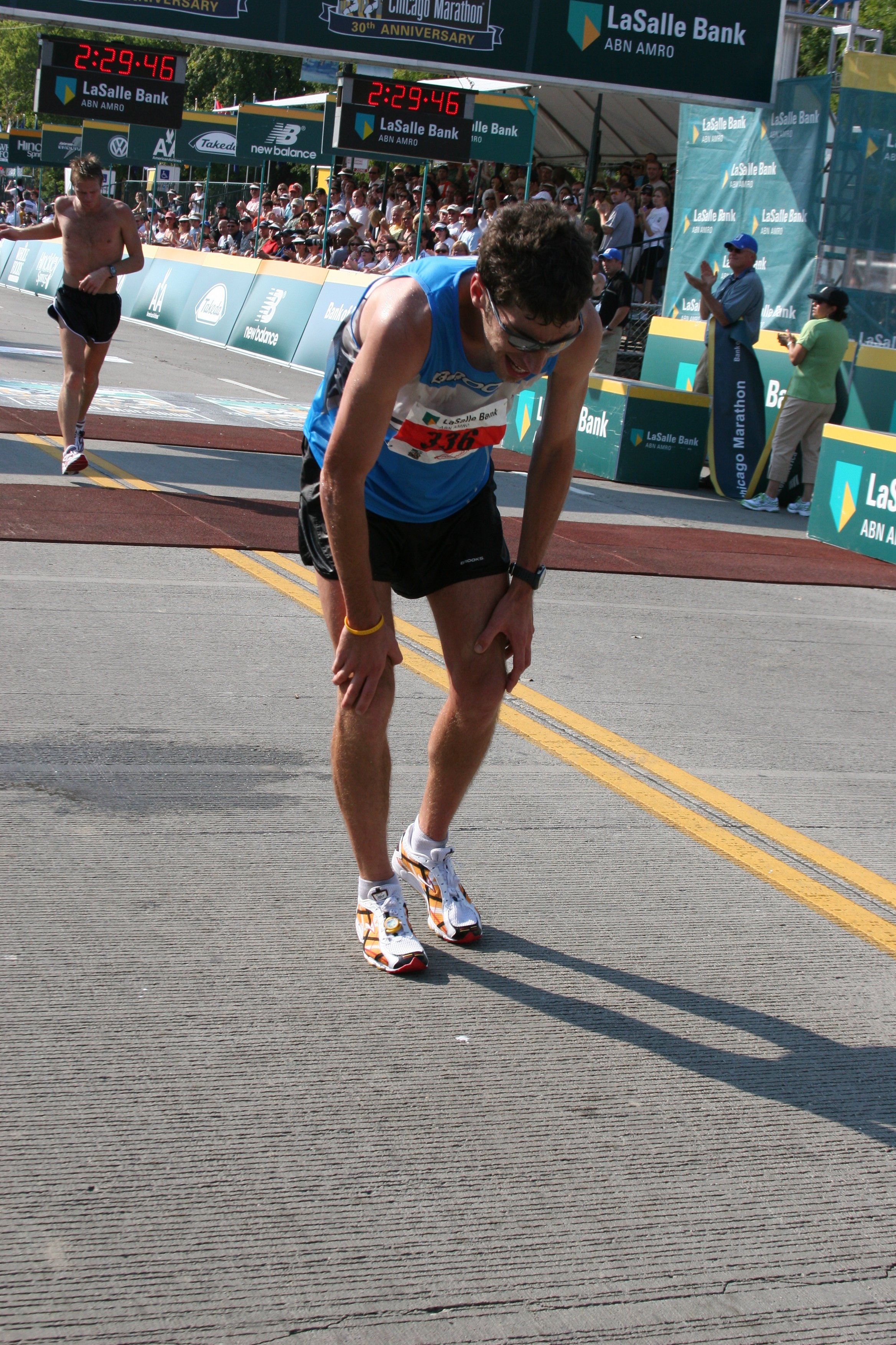 A male marathon runner in a blue tank top and black shorts, bent over and exhausted, crossing the finish line of a race, with another runner behind him near the crowd and banners, some spectators, and officials clapping and cheering.