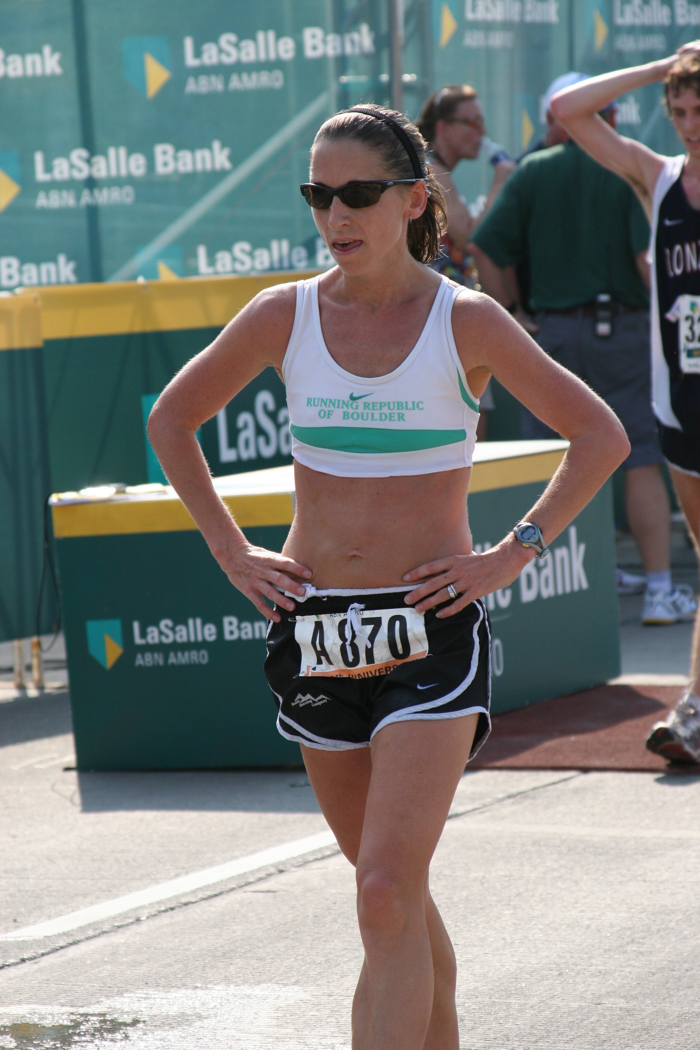 A female marathon runner wearing sunglasses, a white tank top with green text, black shorts, and a race bib, running on a track near a LaSalle Bank sponsored area during daytime.