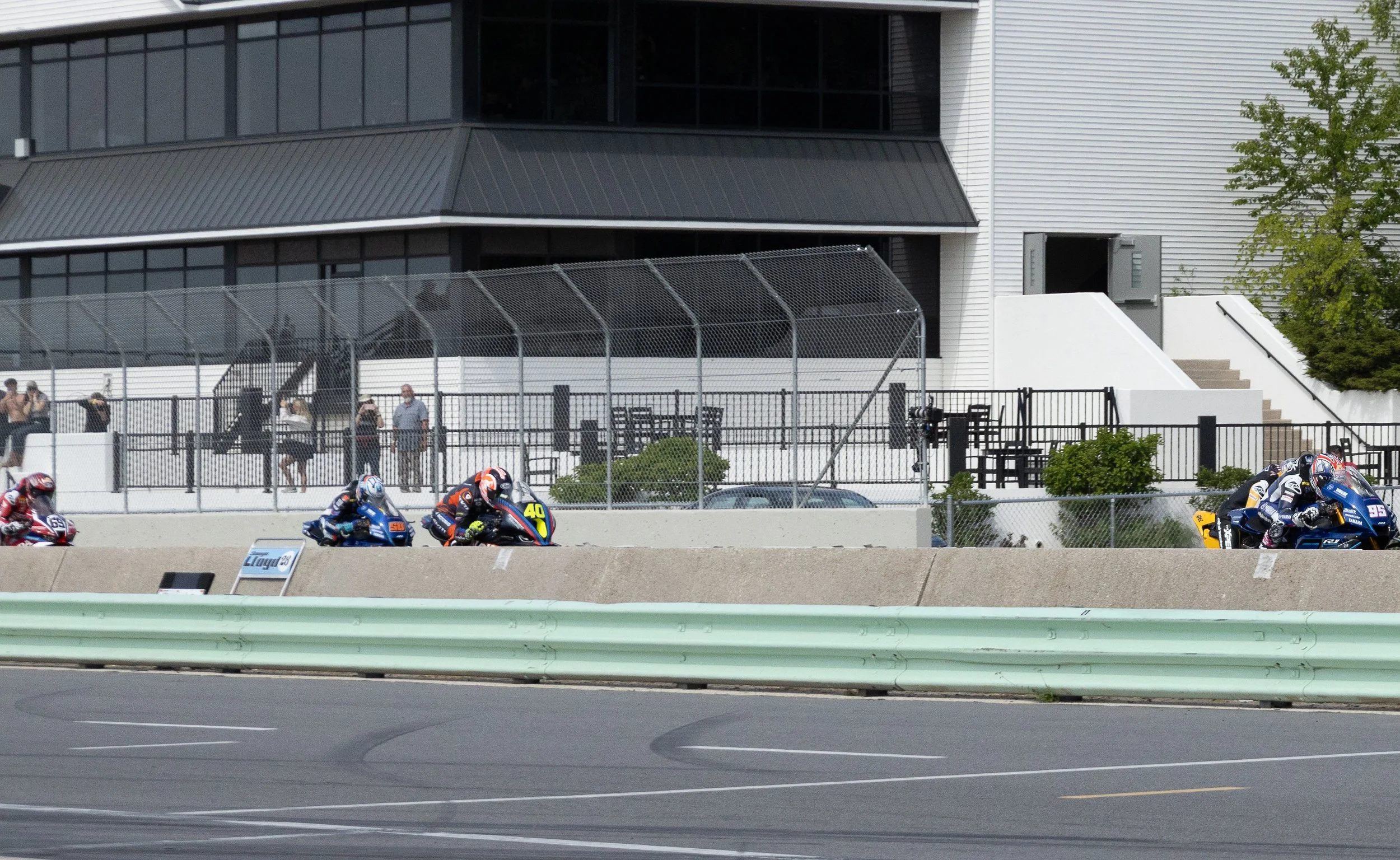 Motorcycle racers competing on a race track with a large modern building and green tree in the background.