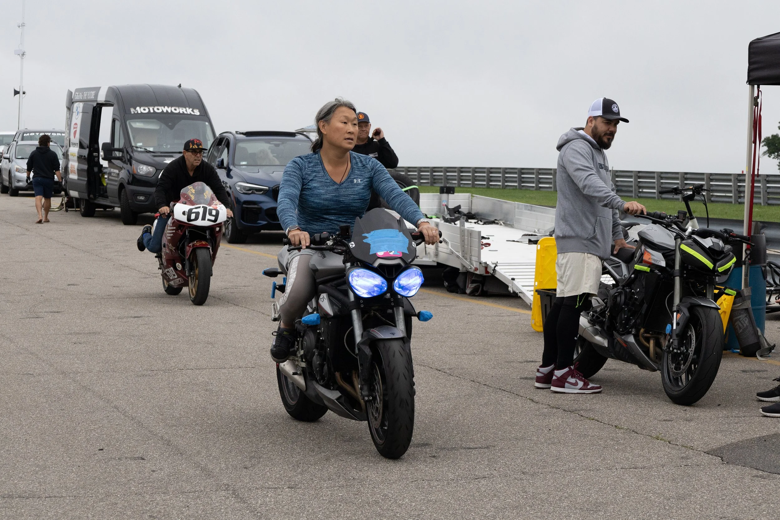 Multiple people preparing for a race at a racetrack, with motorcycles, vans, and cars in the background, on a cloudy day.
