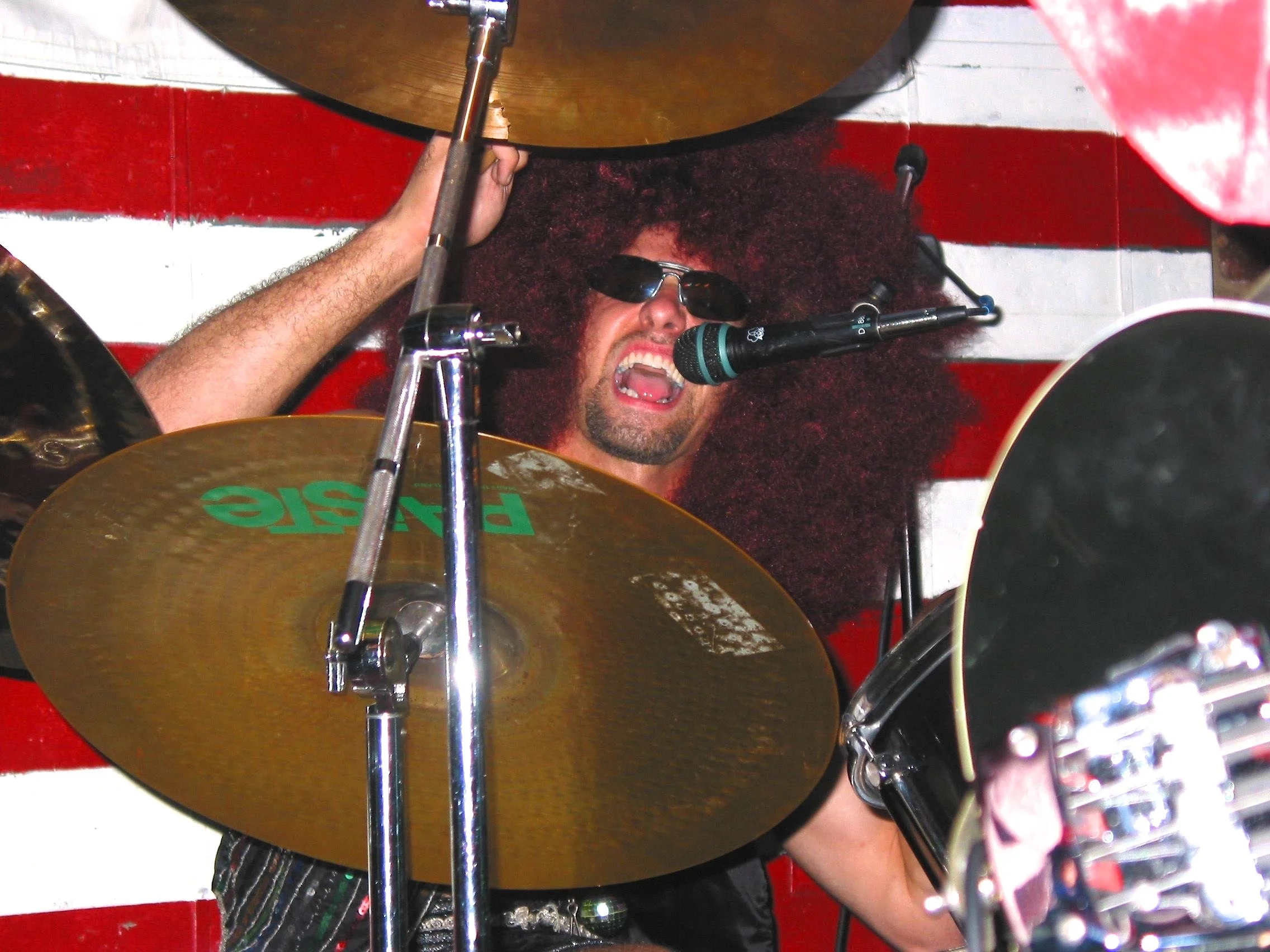 A man with long curly hair, sunglasses, and a beard singing into a microphone while playing drums, with a red and white striped background.