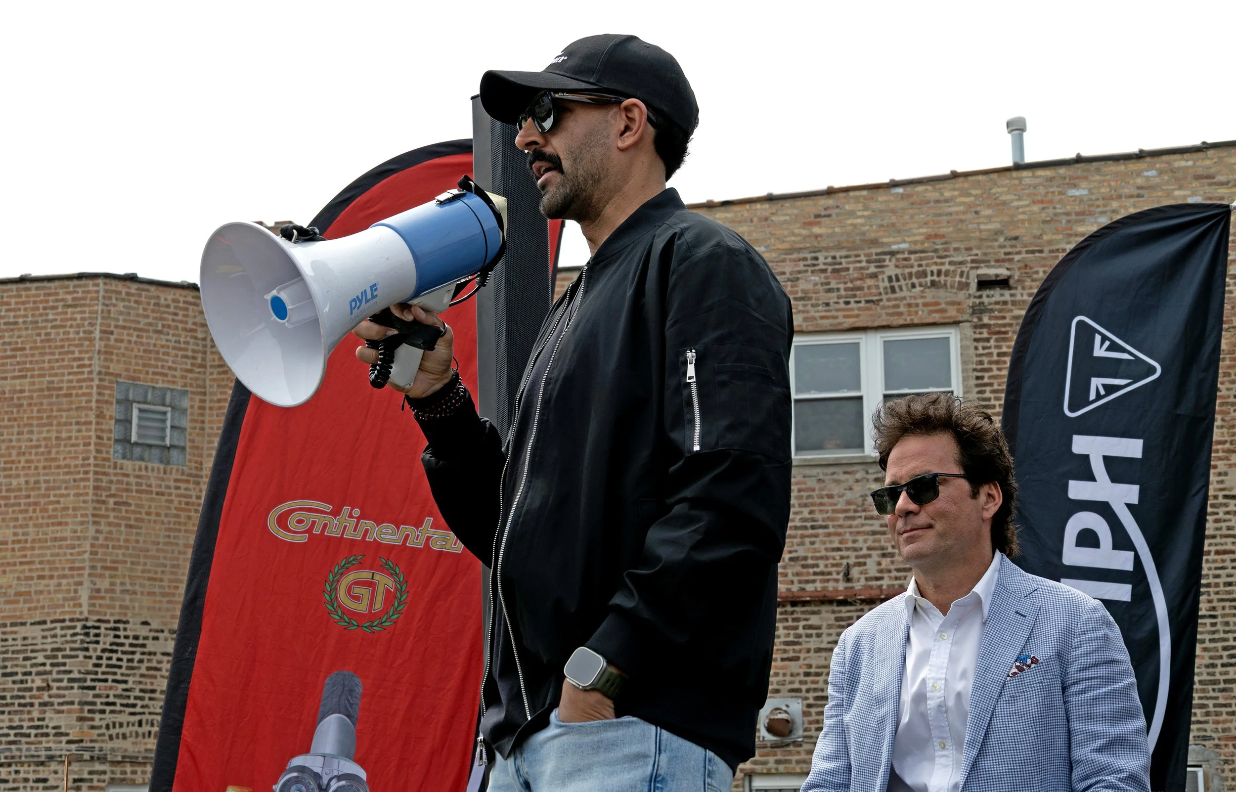A man wearing black sunglasses, a black cap, and a black jacket holding a megaphone speaks at an outdoor event. Two other men, one wearing sunglasses and a checkered blazer and the other with glasses and a shirt, stand nearby. There are flags and ban