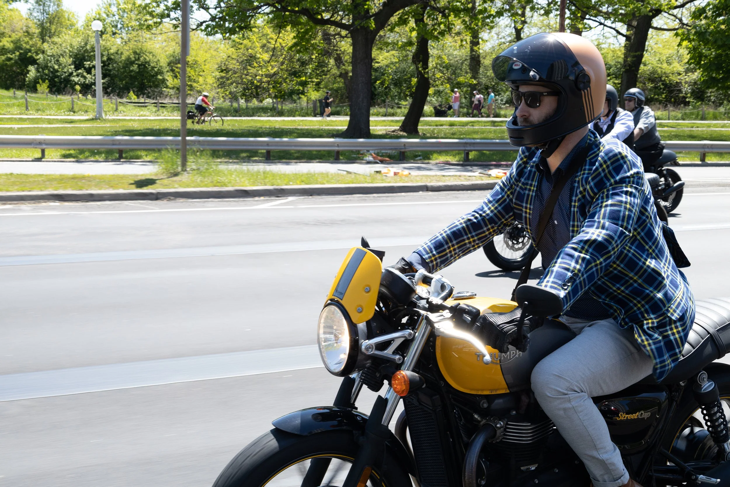 A man riding a yellow and black motorcycle on a city street during the daytime, with other motorcyclists, pedestrians, and trees in the background.