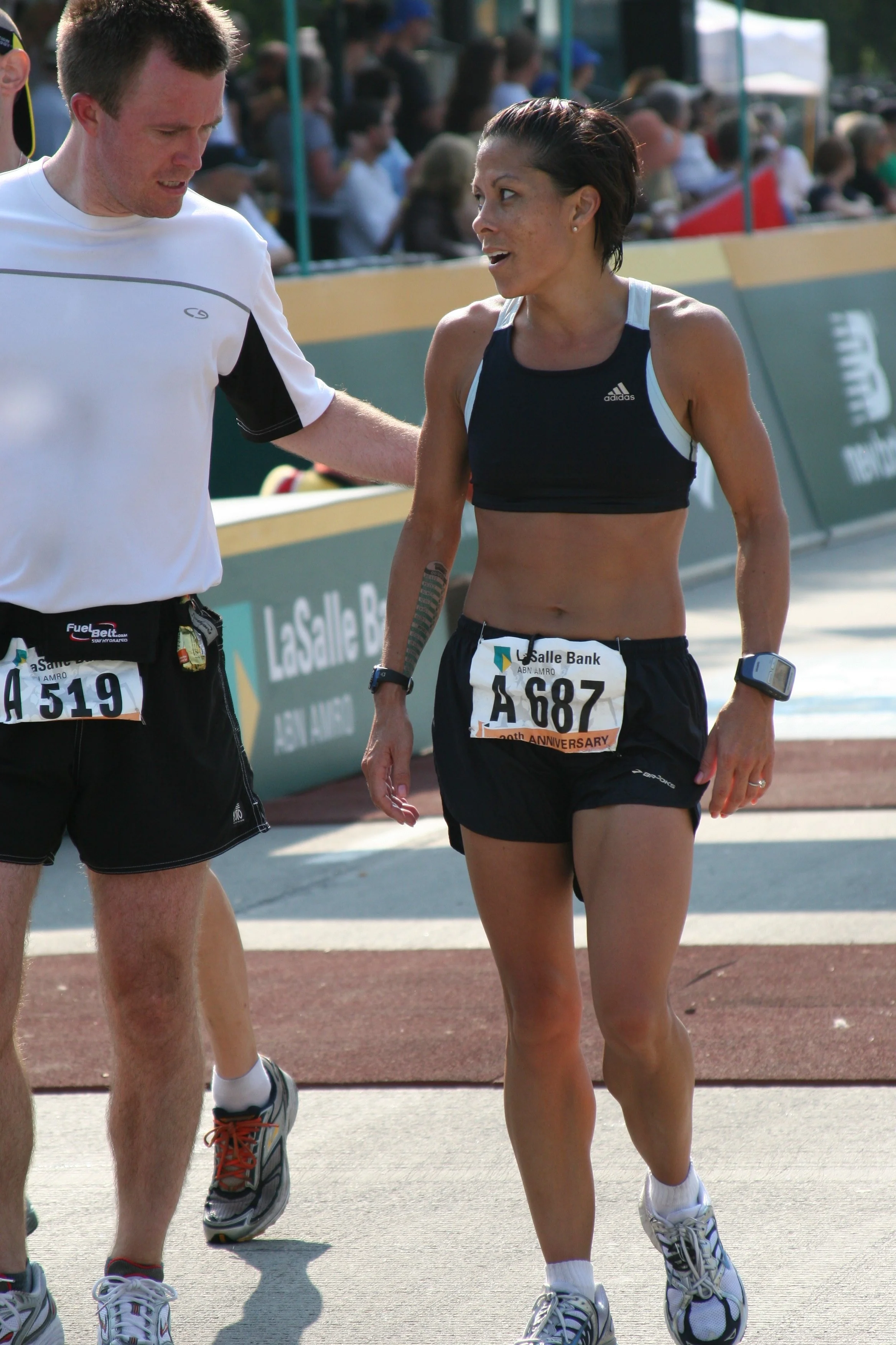 A woman in athletic wear and a man talking during a race, with a crowd and banners in the background.