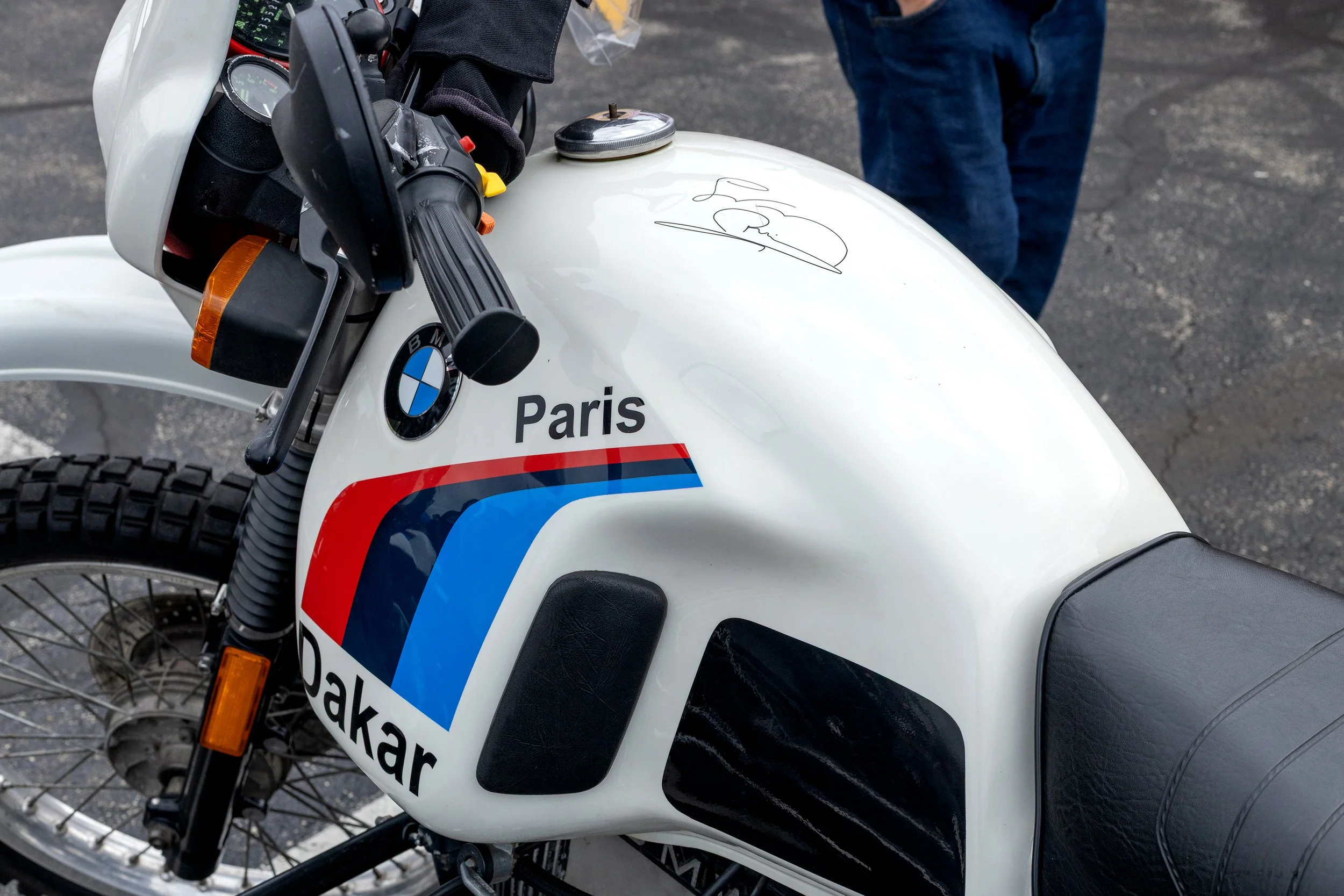 Close-up of a white BMW motorcycle with a signature on the fuel tank, a sign with 'Paris' and a logo that includes red, white, and blue stripes, and a sign that reads 'bakar'. A person's legs in blue jeans are visible in the background, standing on a