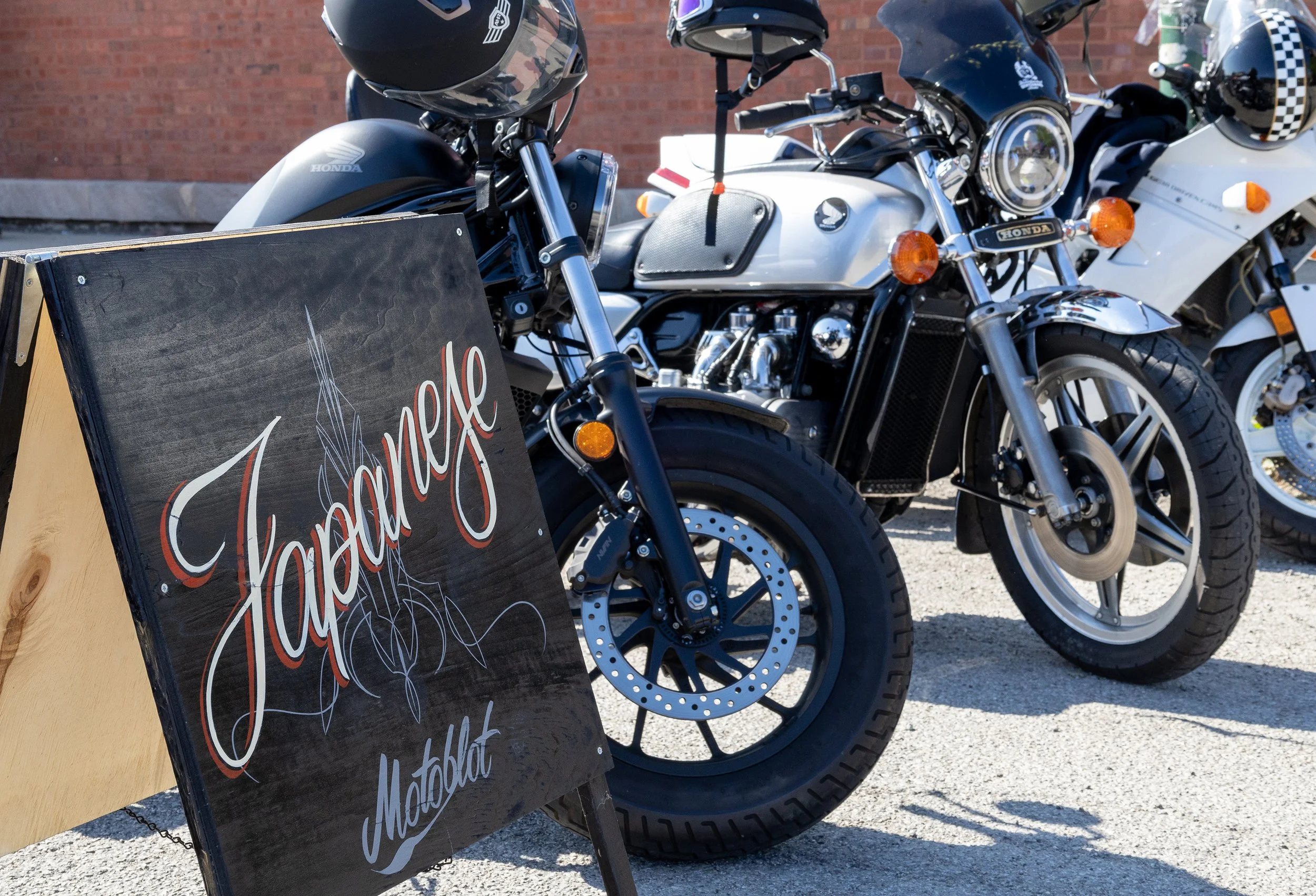 Multiple motorcycles parked outdoors next to a black chalkboard sign with red and white cursive lettering, a brick wall in the background.