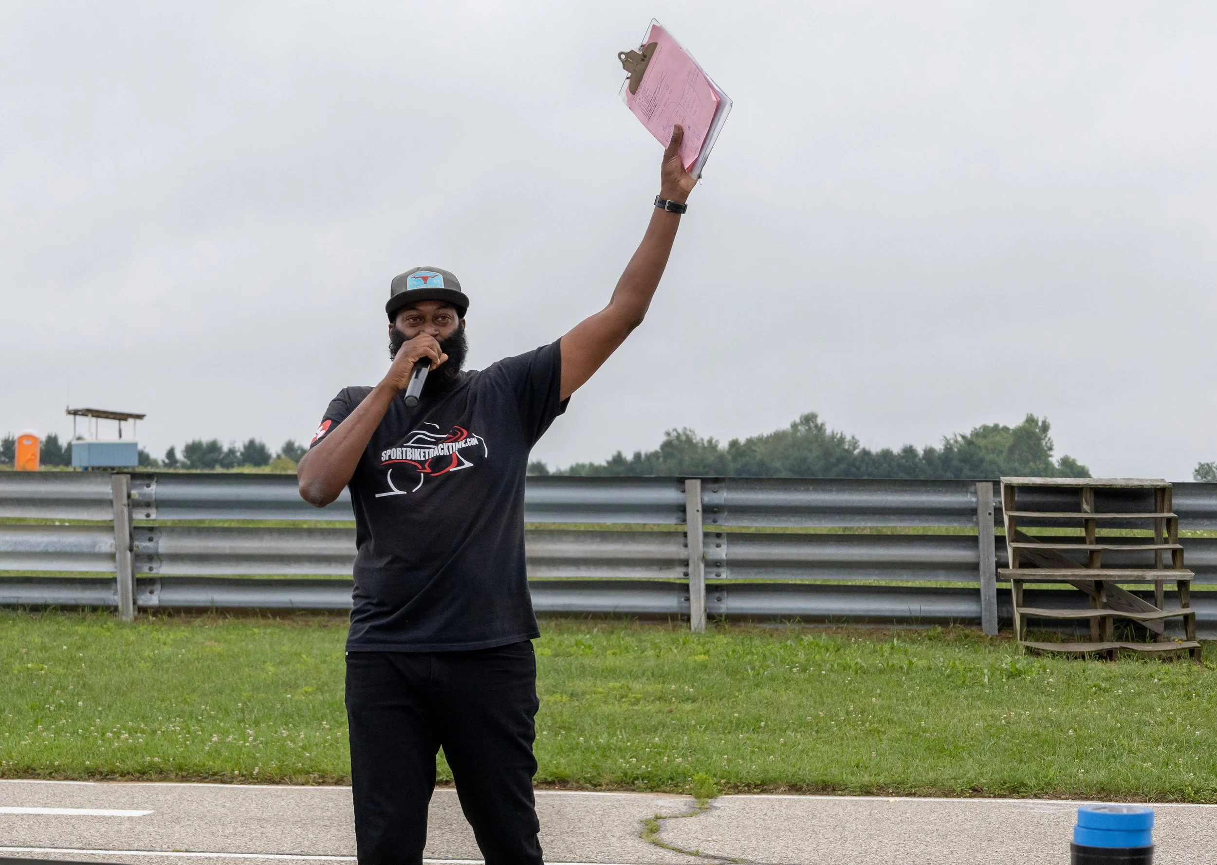 A man holding a clipboard in the air while speaking into a microphone at an outdoor event near a racetrack.