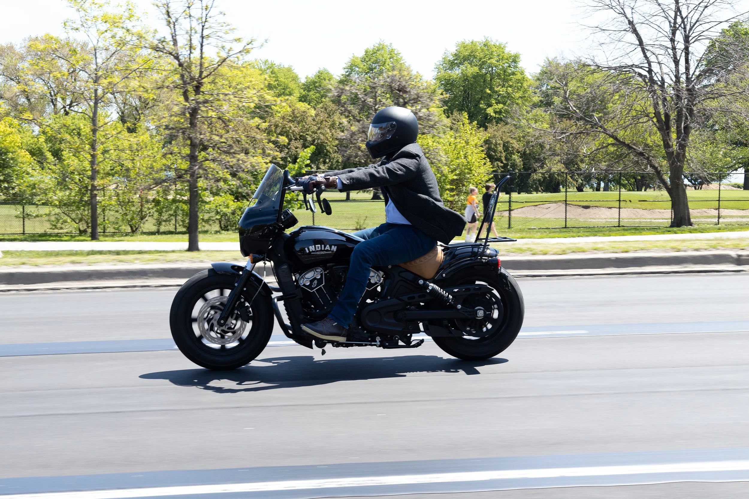 A man riding a black motorcycle on a paved road in a park with green trees and a fence in the background, wearing a helmet and a black jacket.