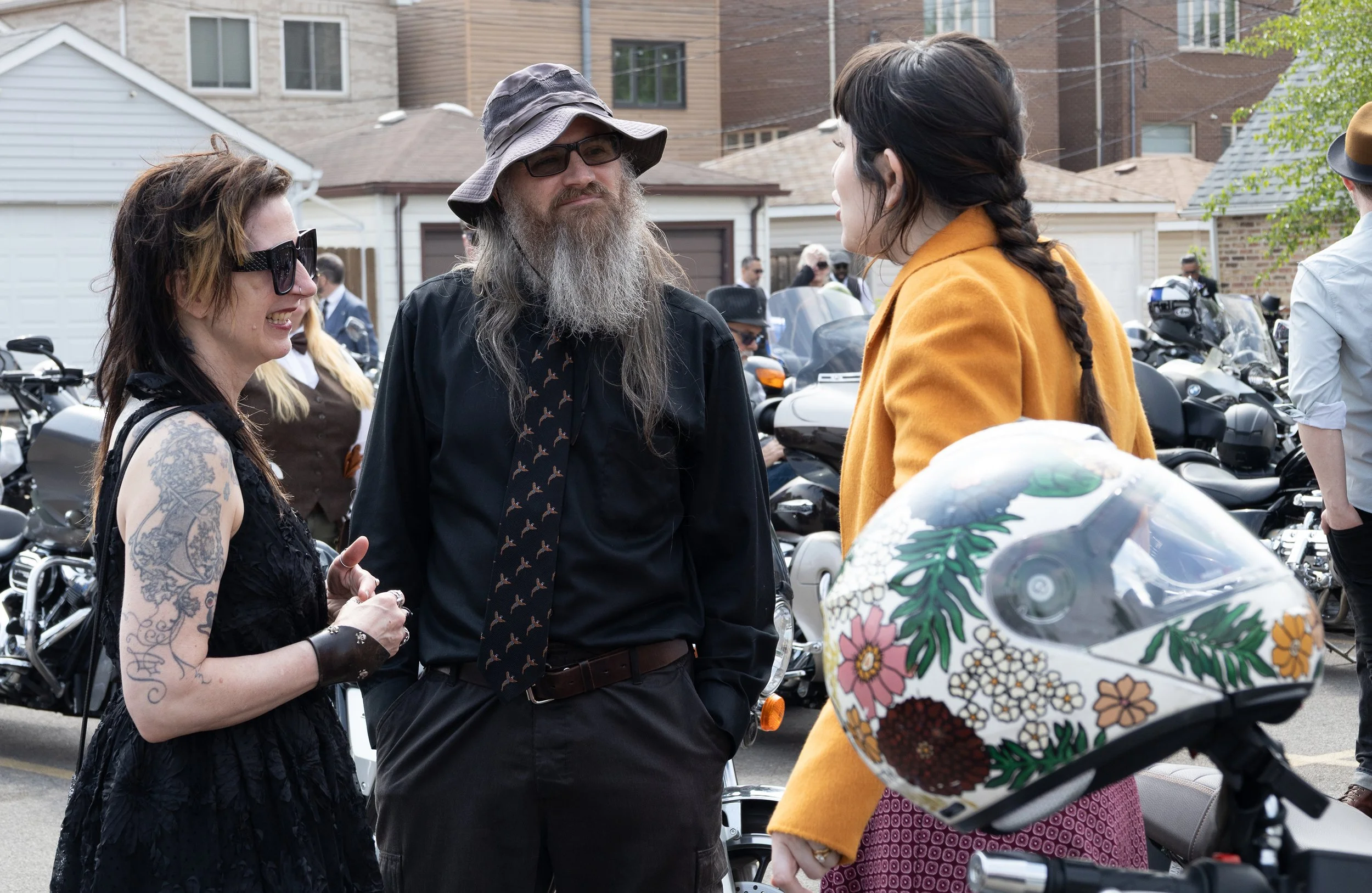 Three people are talking on a street with motorbikes parked behind them. One woman with tattoos and sunglasses, a man with a long beard and hat, and another woman with a braid and a mustard-colored jacket are engaged in conversation.
