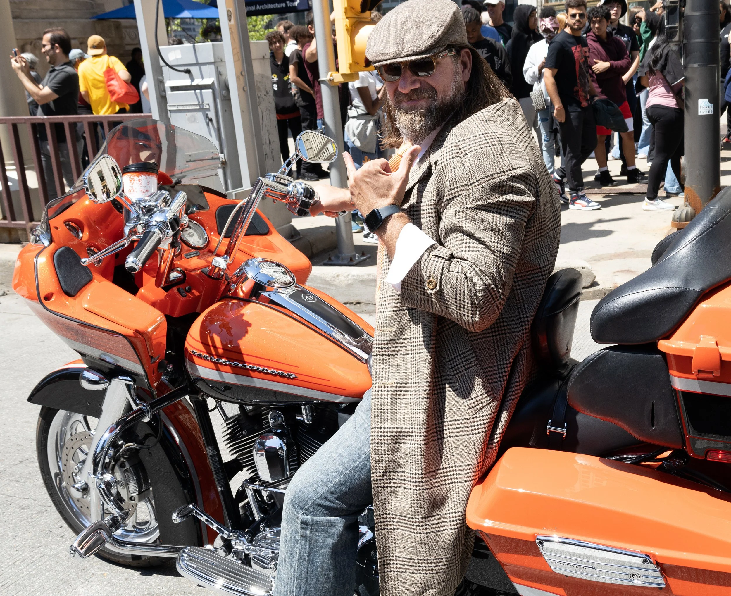 A man with long hair, beard, sunglasses, and a plaid blazer sitting on an orange motorcycle in a crowded outdoor event.