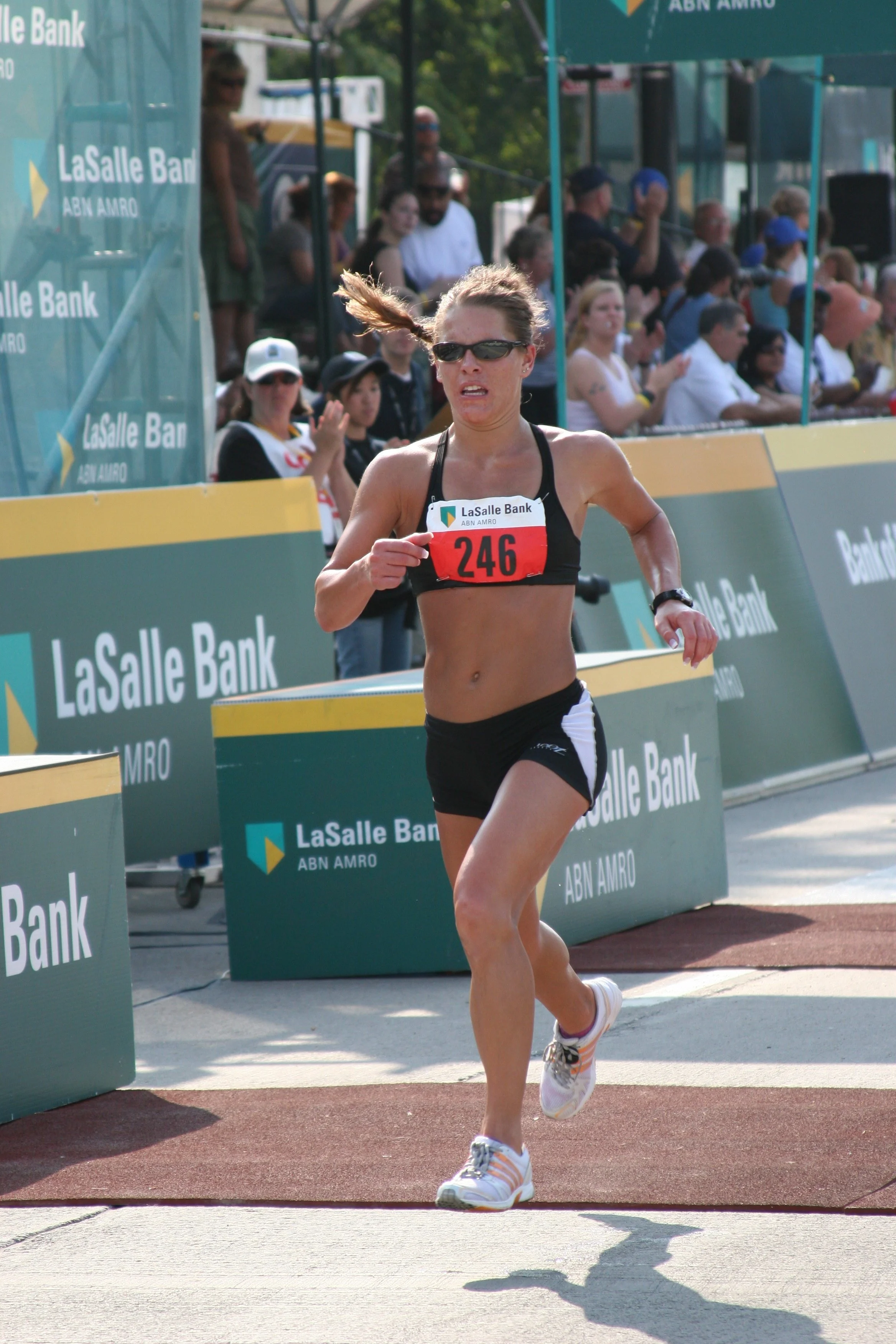 Female athlete wearing sunglasses and a black sports bra with the number 246, running in a race at an outdoor track, with spectators and banners in the background.