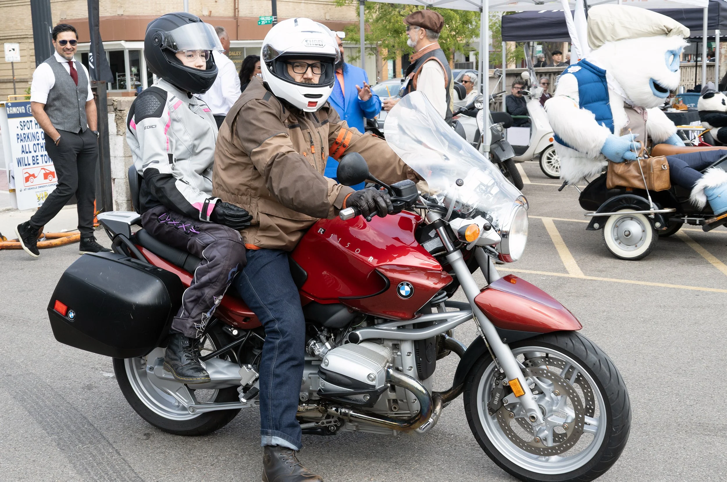 Two people wearing motorcycle helmets riding a red BMW motorcycle, with a crowd and tents in the background during an outdoor event.