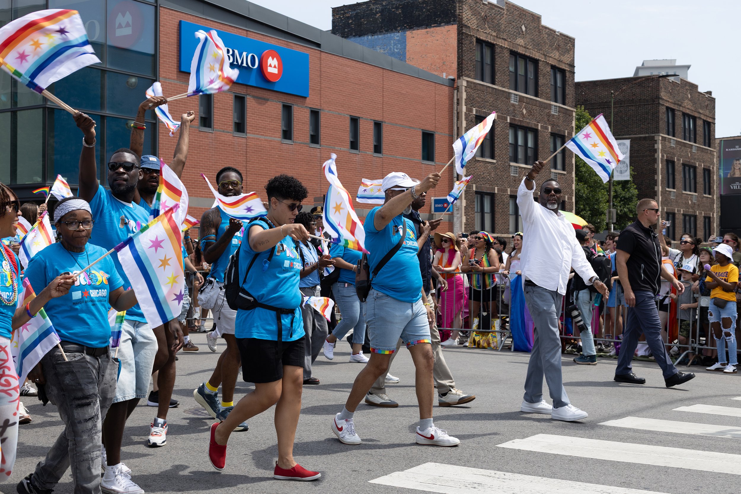 A diverse group of people participating in a pride parade, walking across a street while waving rainbow and pride flags. The crowd is dressed in colorful clothing, and there are spectators on the sidewalk. Buildings and a BMO bank sign are visible in