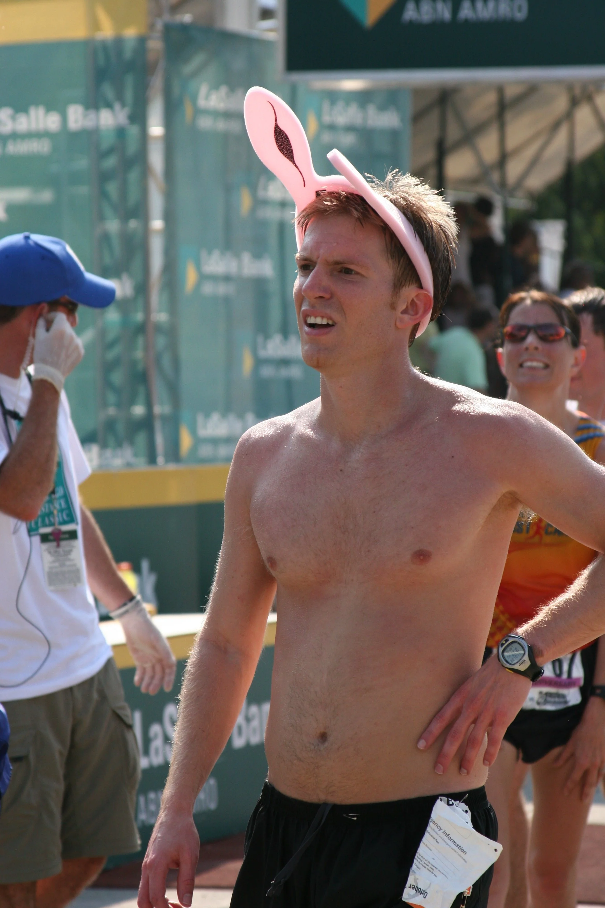 A shirtless man with a pink bunny ears headband running at a race event, surrounded by other runners and event staff.