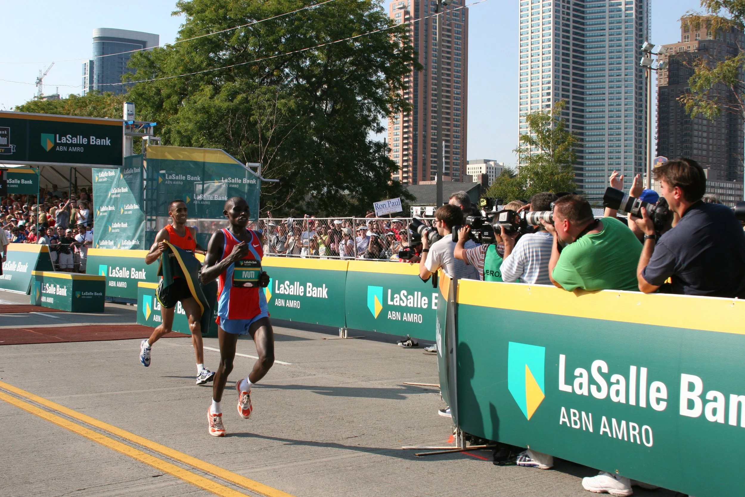Marathon runners crossing the finish line at a race sponsored by LaSalle Bank, with spectators and photographers taking pictures nearby, in an urban area with high-rise buildings behind.