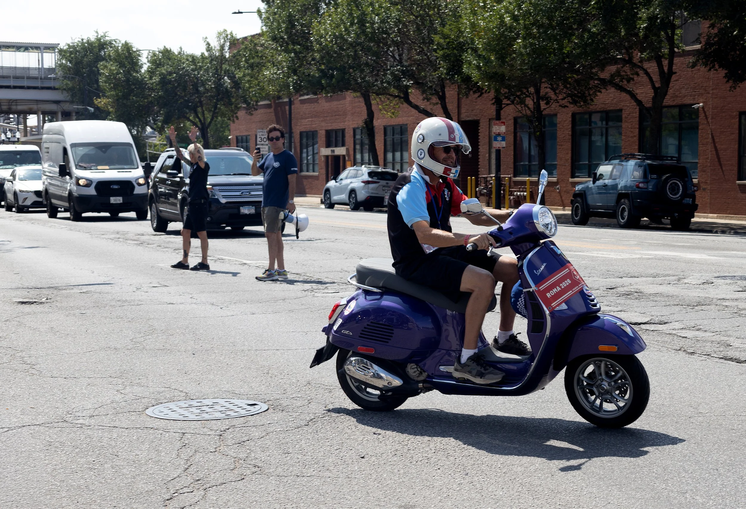 A man riding a purple Vespa scooter with a helmet, sunglasses, and a 'Roma 2026' sign. In the background, a woman with blonde hair and a man with dark hair and sunglasses stand on a city street, waving and taking a photo. Several parked cars and a br