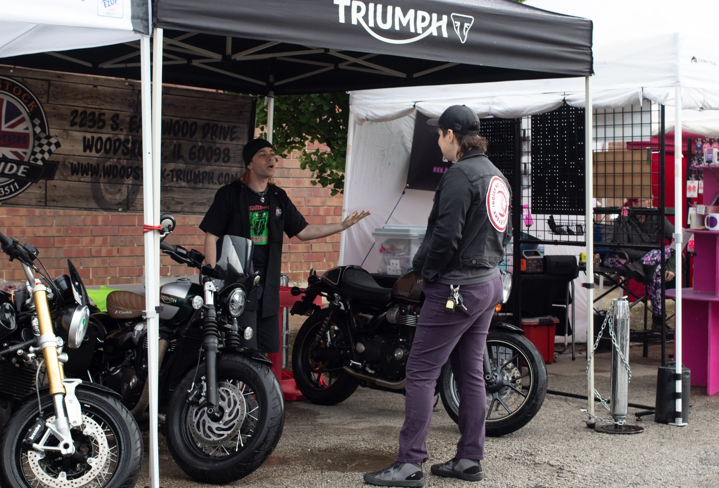A man and a woman talking at an outdoor motorcycle booth. The booth has a black canopy with 'Triumph' written on it, and several motorcycles are on display. The man is wearing a black jacket and a black cap, while the woman has a black jacket with a 