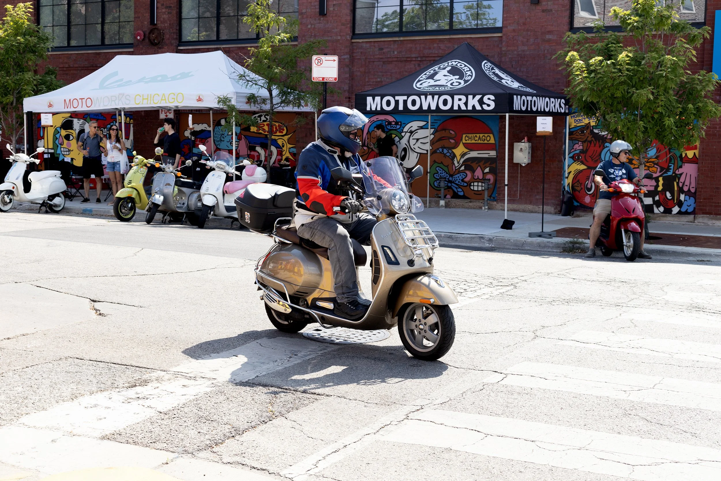 A motorcyclist wearing a blue helmet and jacket riding a beige scooter on a city street. Two tents with colorful artwork and people underneath are in the background, along with parked scooters and a brick building.