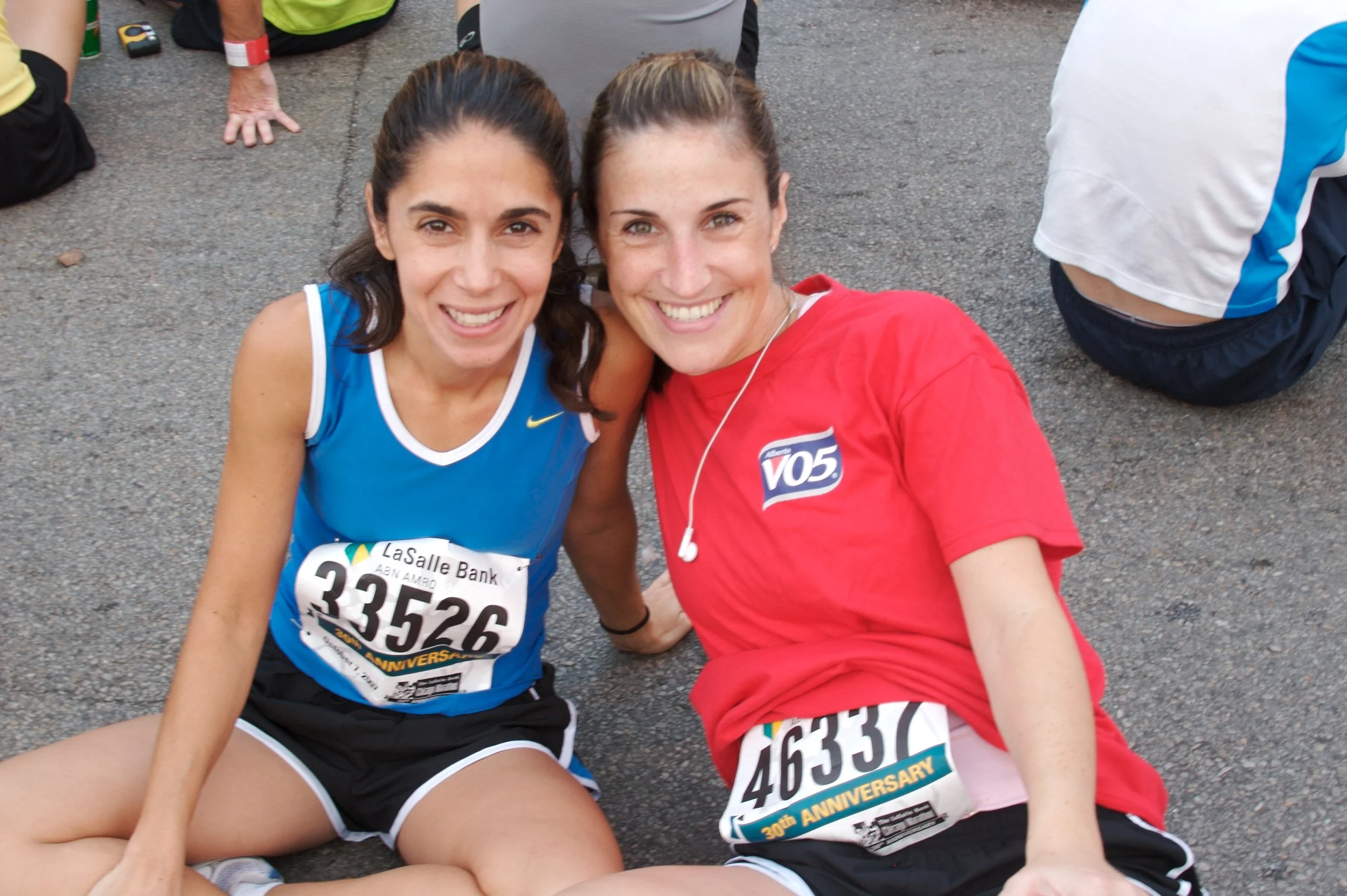 Two women are sitting on the ground during a race or marathon, smiling at the camera. They are wearing athletic clothes, with race bibs attached, and appear to have just finished running. One woman has dark hair tied back and is wearing a blue top, w