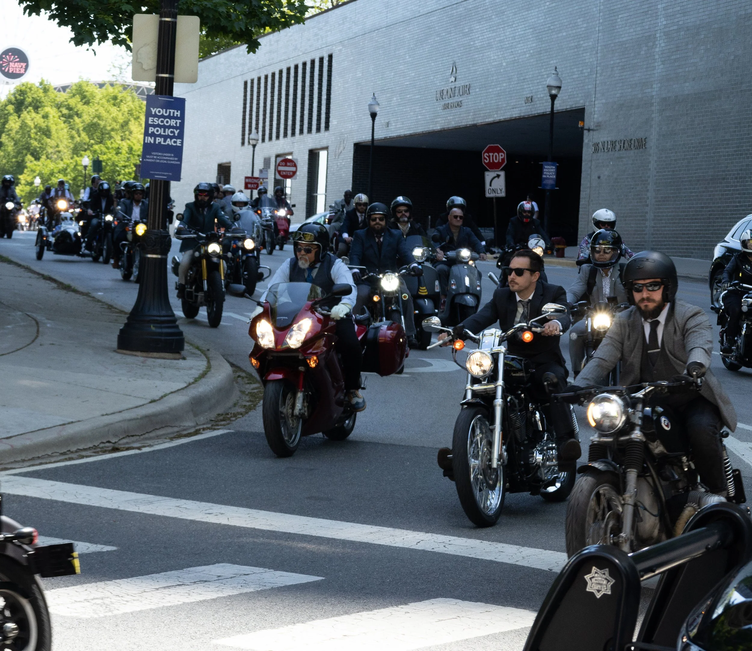 A group of motorcyclists riding together on a city street, with some wearing helmets and sunglasses, next to a building and street signs.