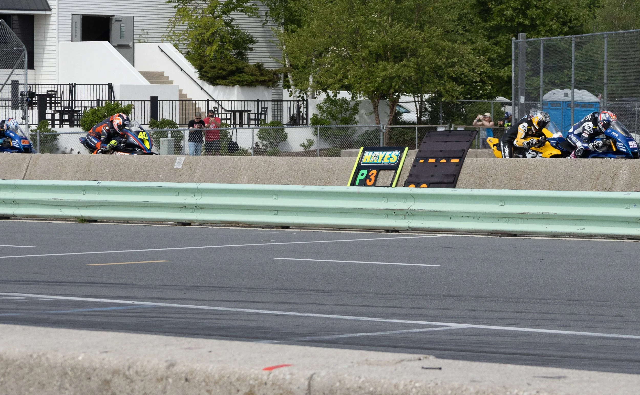 Motorcycle racers in racing suits and helmets riding on a racetrack with spectators behind a fence, and a sign indicating the race type.
